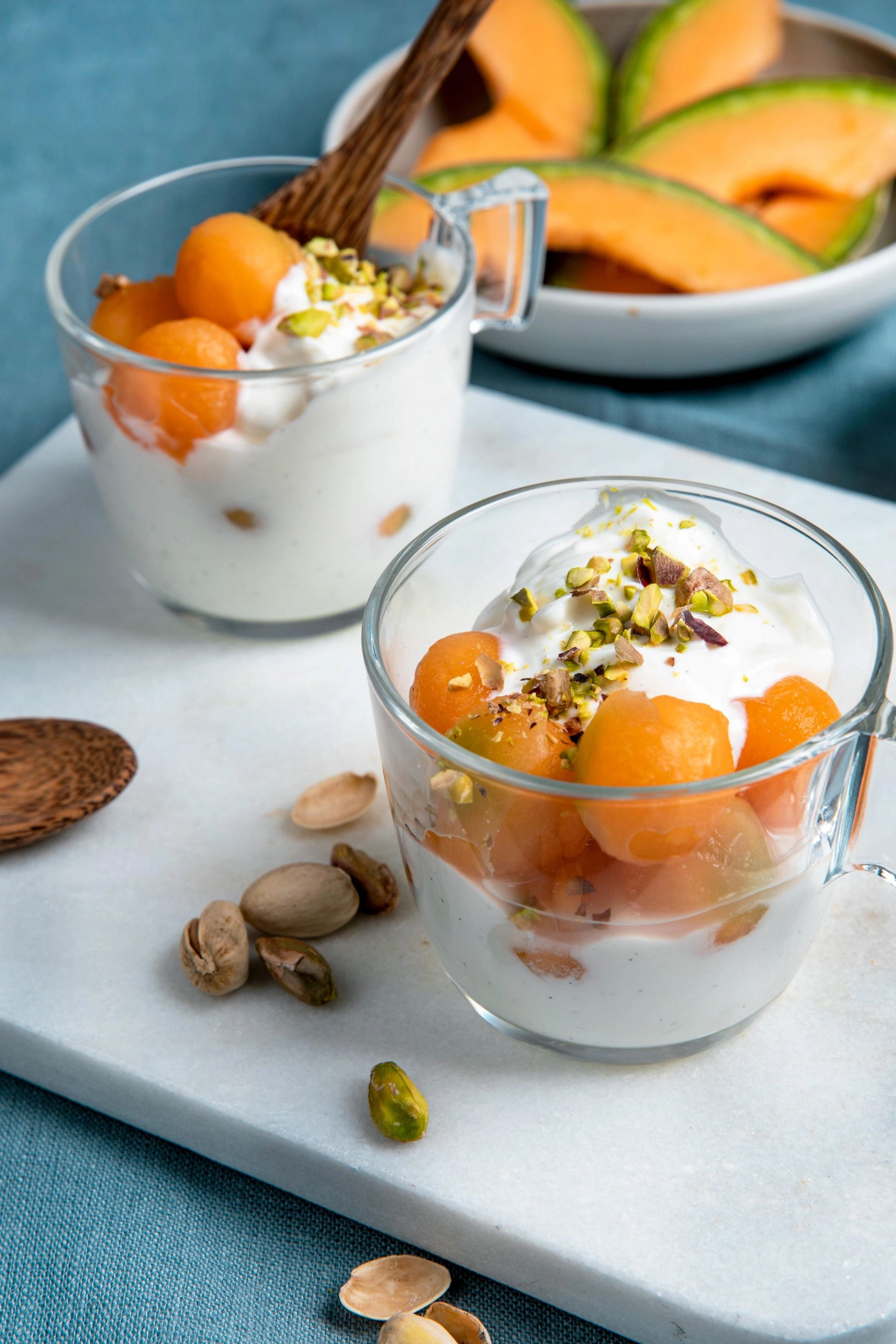 To glasskopper fylt med yoghurt toppet med melon‑kuler og hakkede pistasjnøtter står på et brett, med melonskiver liggende i en skål i bakgrunnen.