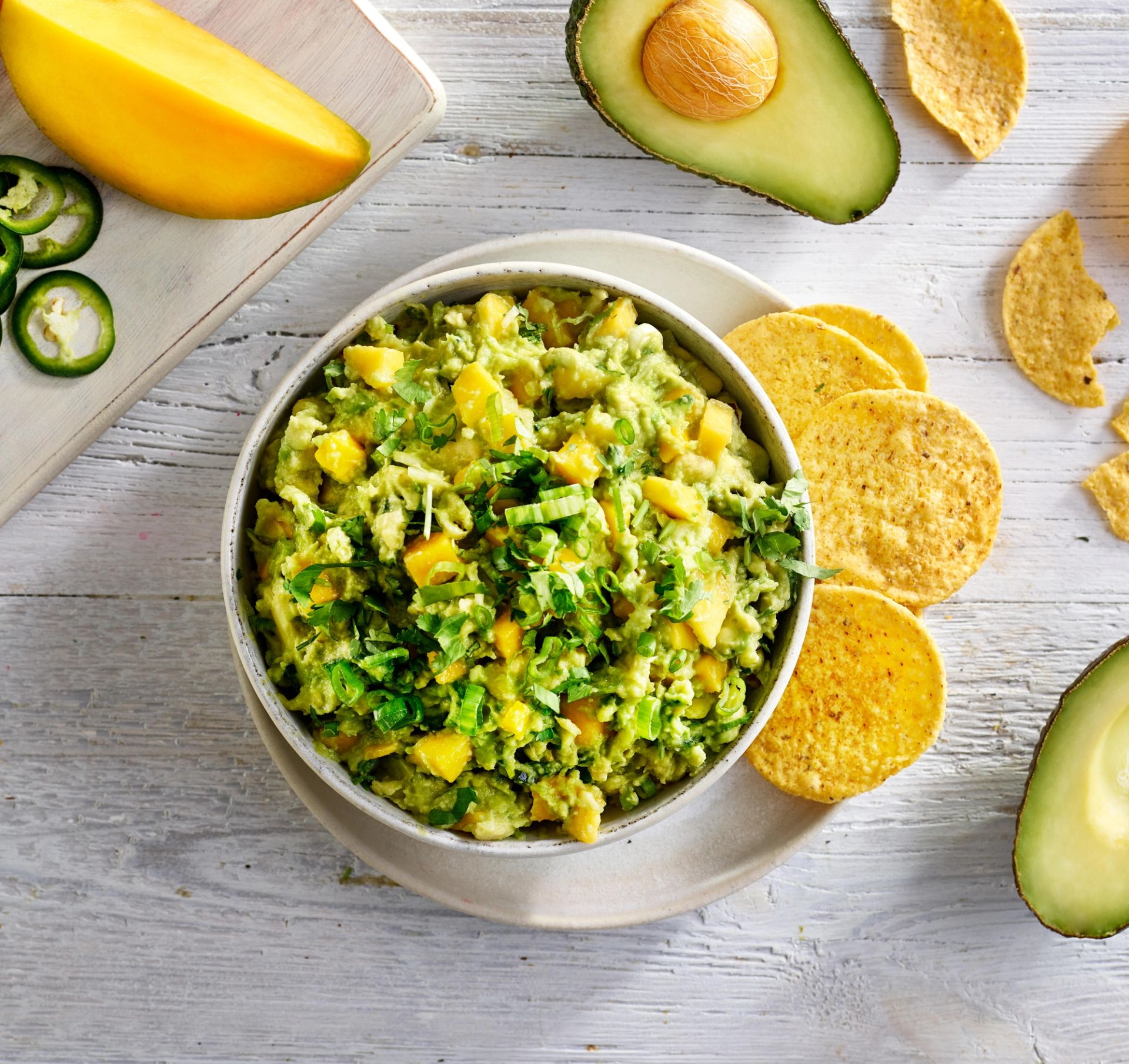 En skål med chunky guacamole med mango og frisk koriander, omgitt av tortillachips, avokadohalvdeler, jalapénoringer og mango på et lyst trebord.