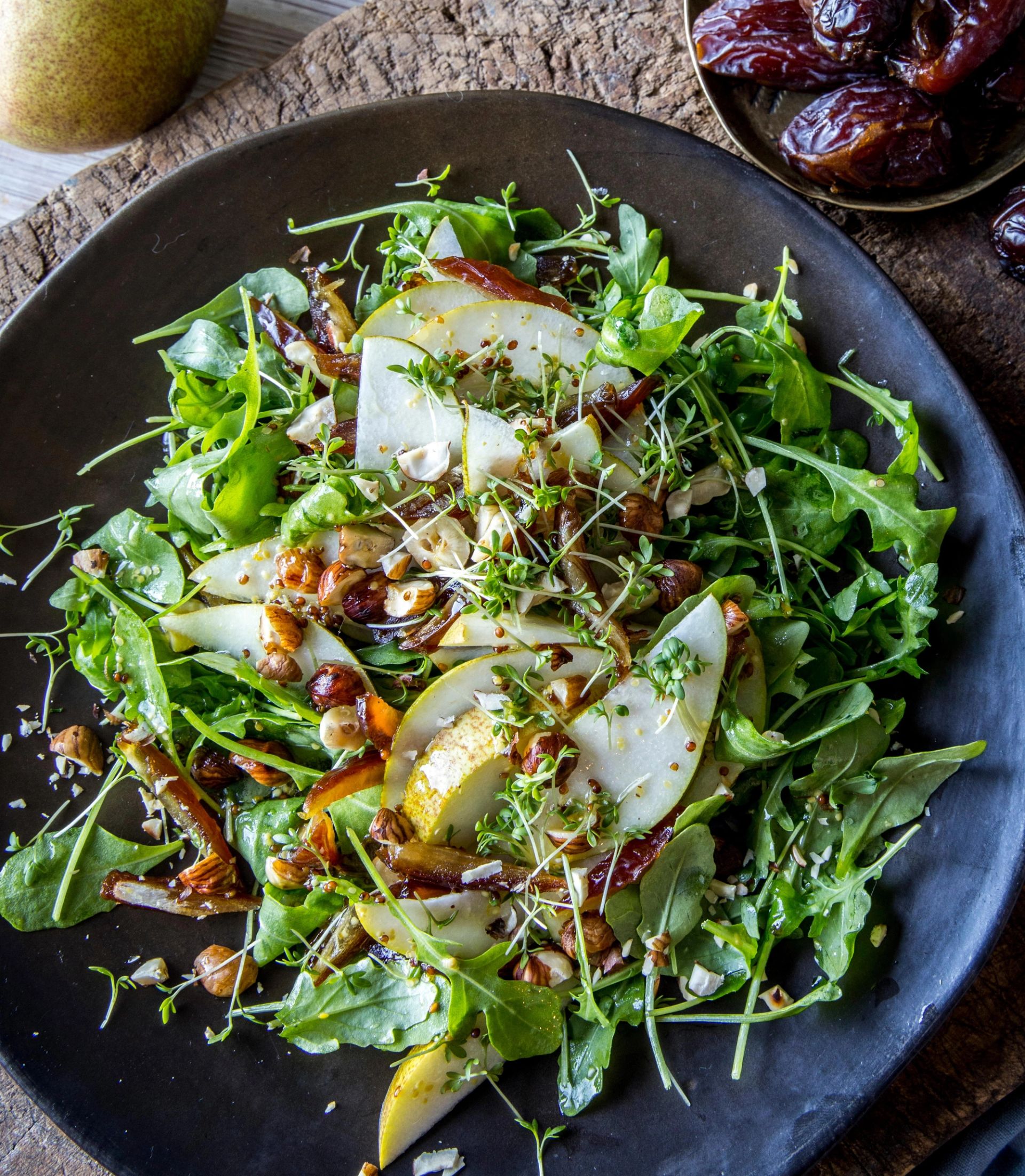 Salat i mørk bolle sett ovenfra, med grønne blader, skiver av pære, karamellisert løk, nøtter og spirer, servert på rustikk overflate med dadler i skål ved siden av.