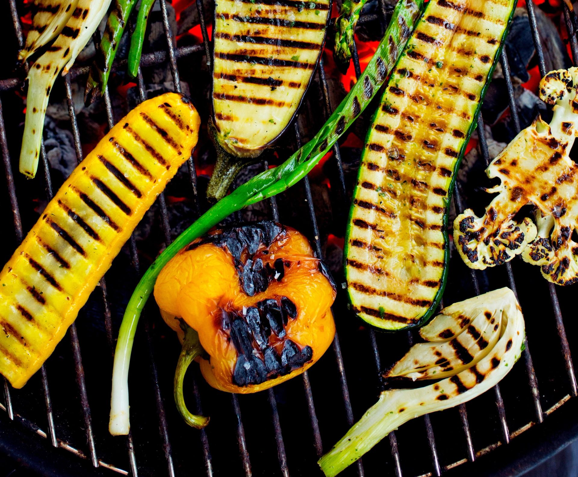 Grillede grønnsaker ligger på en grillrist over glødende kull. På risten ligger blant annet skiver av squash med grillstriper, asparges, en gul paprika med svidde kanter, og biter av rødløk og hvit løk.
