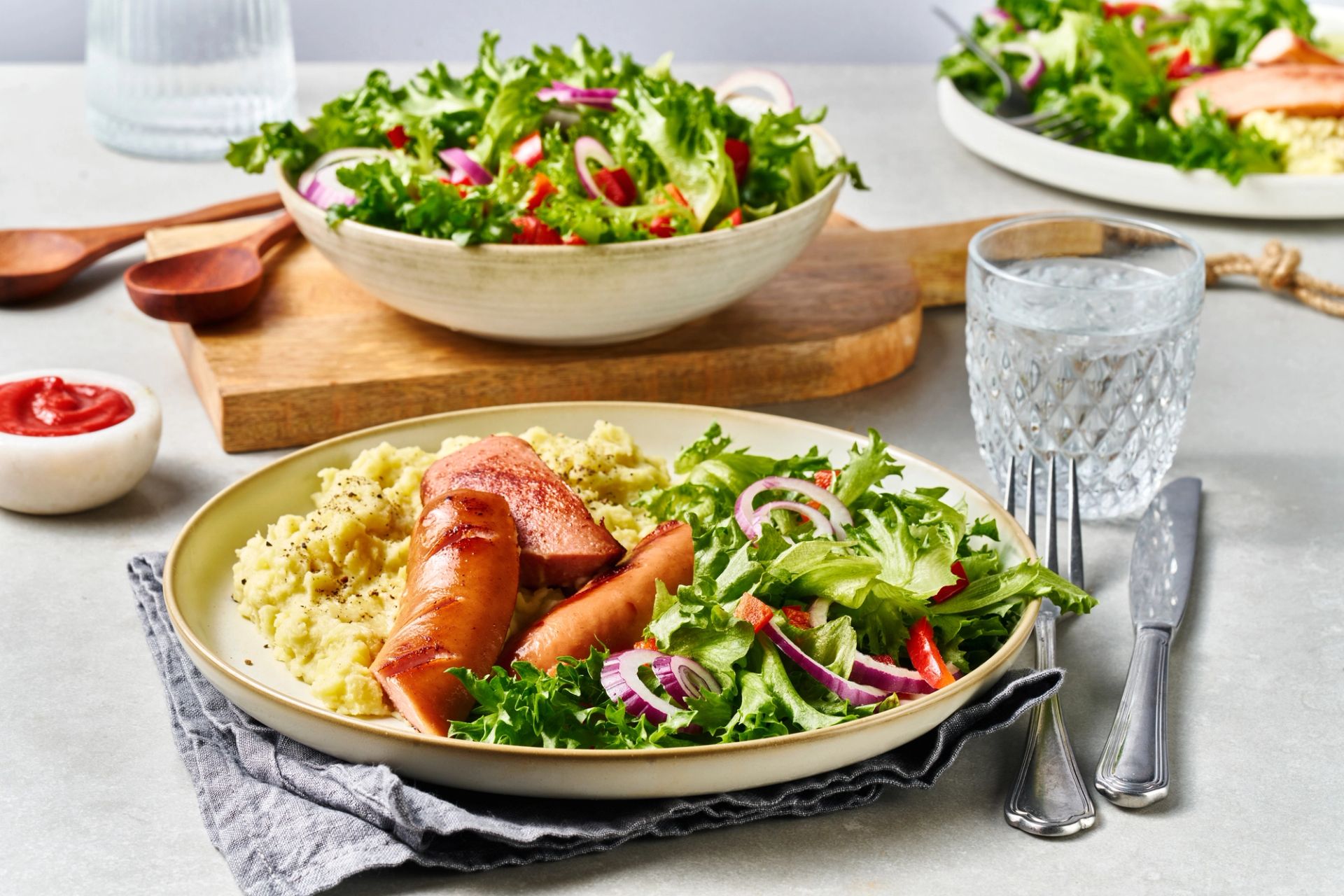 En fristende middagstallerken med stekte kjøttpølse, kremet potetstappe og en frisk blandet salat med rødløk og paprika, servert på et lyst bord med et glass vann ved siden av.