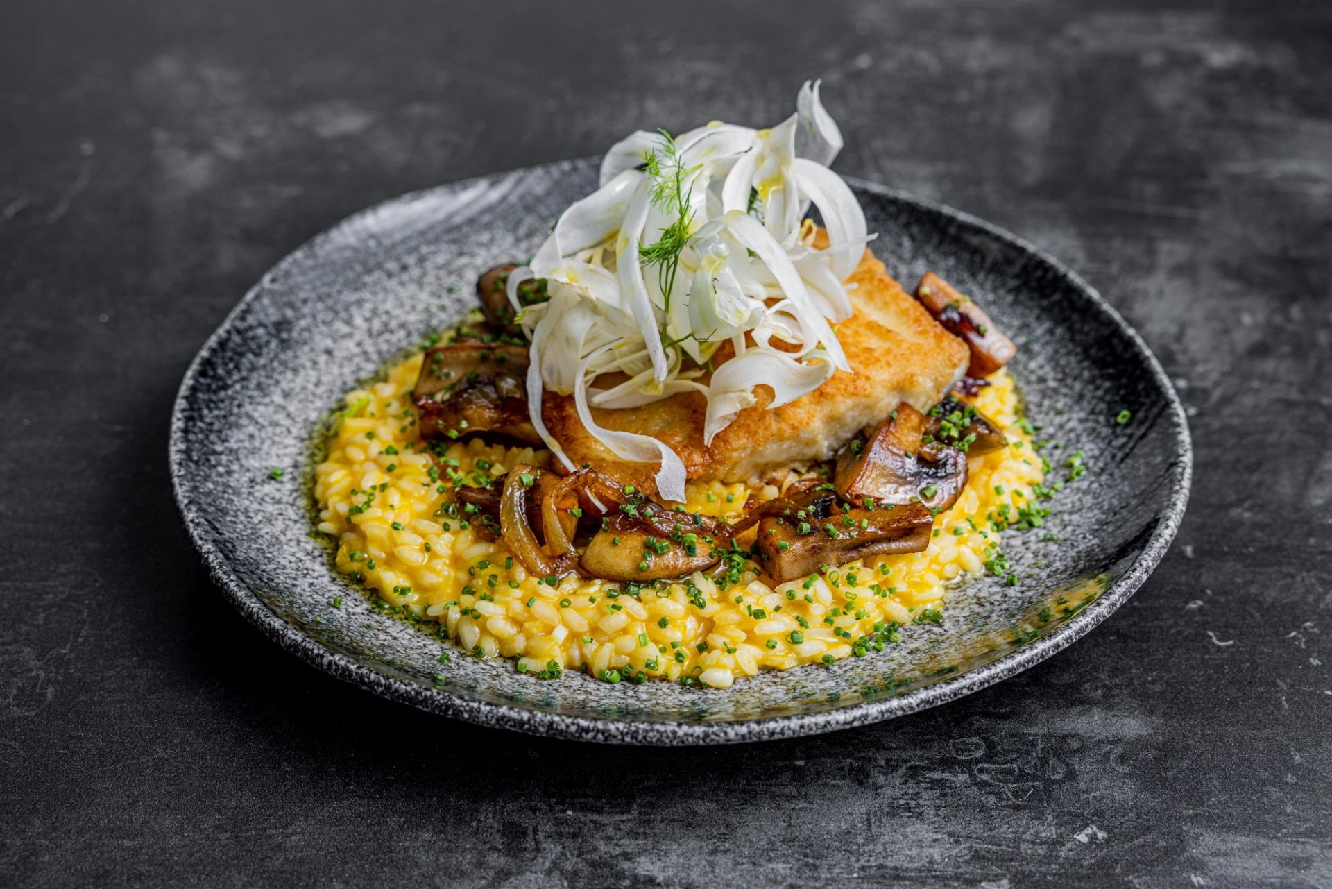 Pannestekt fiskefilét servert på kremet byggrynsrisotto med sautert sopp, toppet med tynne skiver rå fennikel og friske urter, anrettet på mørk tallerken.