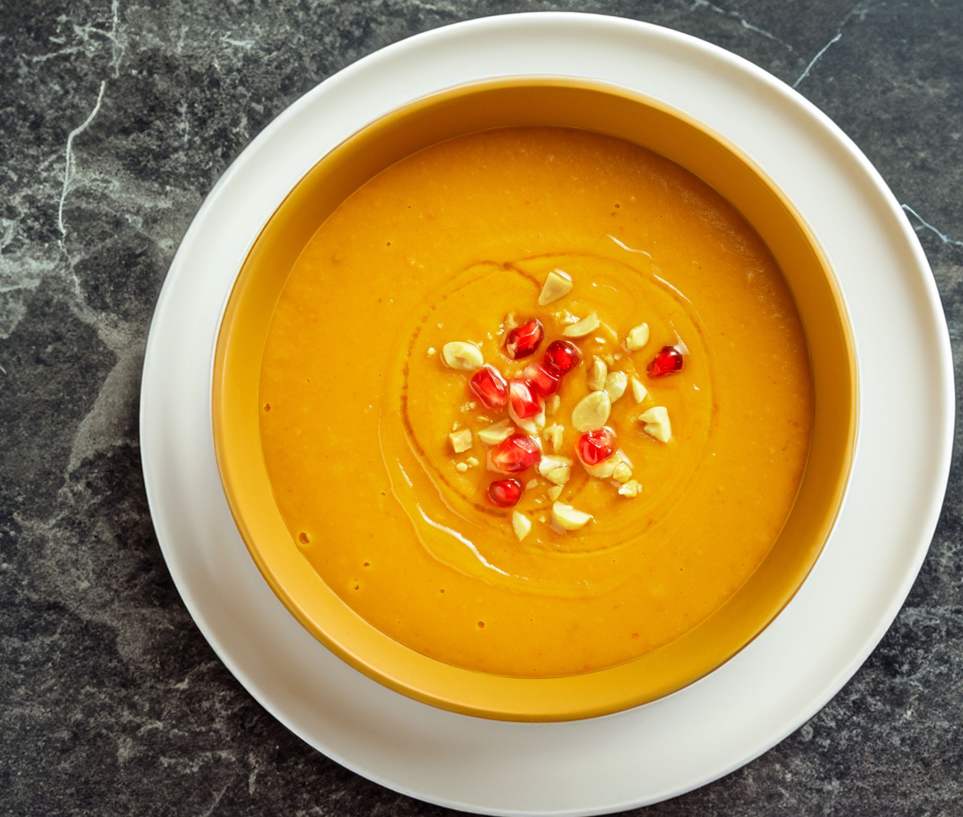 En kremet, glatt søtpotetsuppe i en dyp tallerken, toppet med hakkede nøtter og granateplekjerner, servert på en mørk steinbenkeplate.
