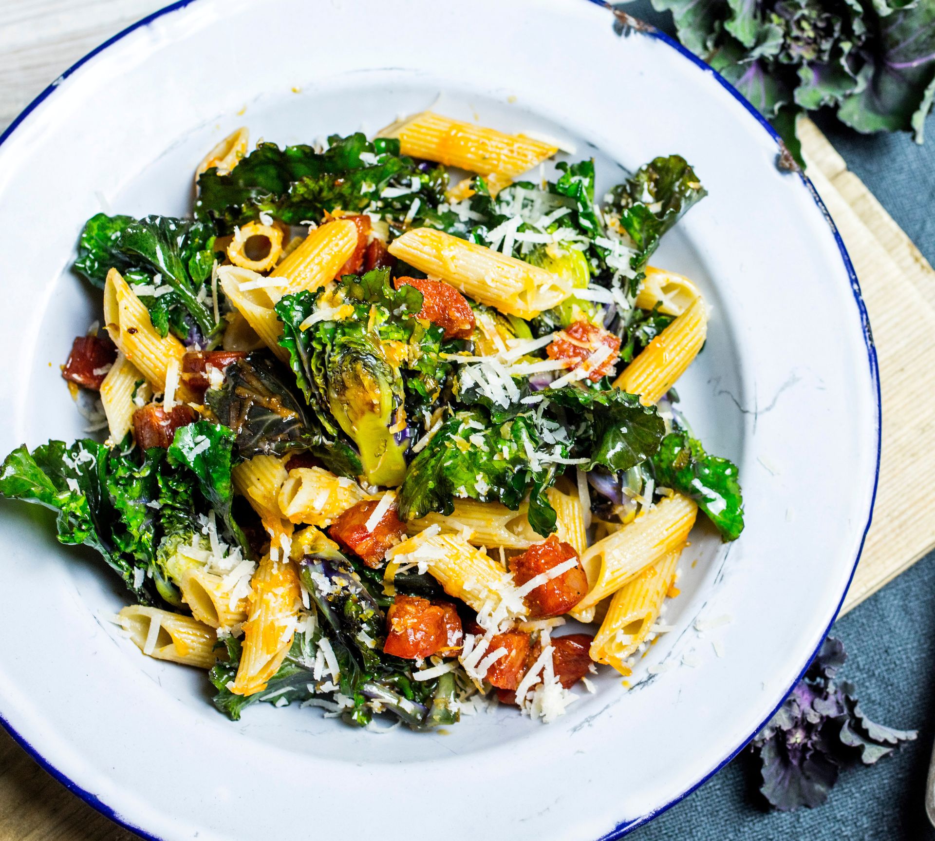 Pastasalat servert i en dyp, hvit tallerken med blå kant. Retten består av pennepasta, rosettkål og chorizo, vendt i olivenolje og toppet med revet ost. De friske grønnsakene og de sterke fargene gir retten et innbydende og rustikt uttrykk.