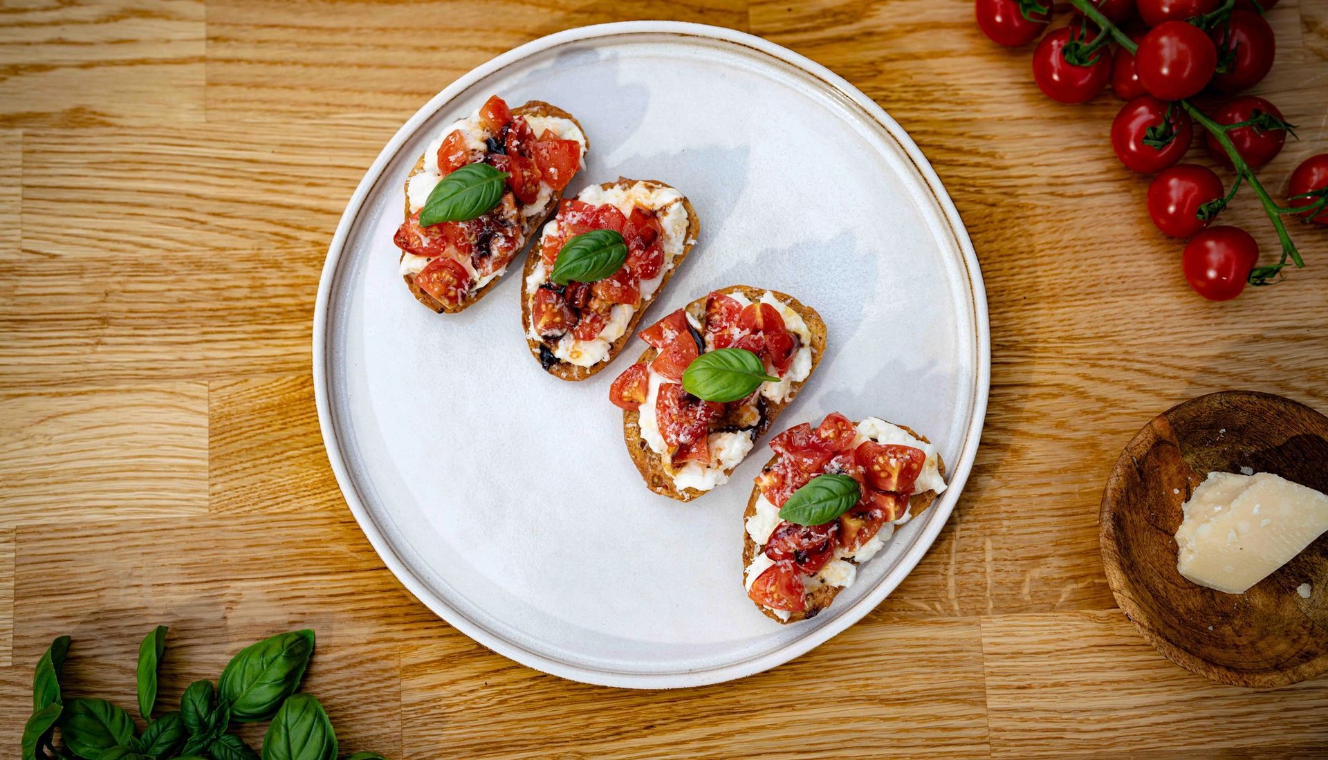 Tallerken med små brød toppet med cherrytomater, friske basilikumblader, mozzarella ost og pinjekjerner. Retten står på en grå overflate, med en gaffel ved siden av og små skåler med røde tomater rundt.