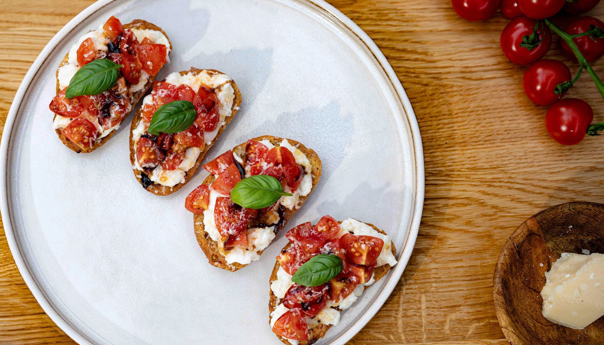 Tallerken med små brød toppet med cherrytomater, friske basilikumblader, mozzarella ost og pinjekjerner. Retten står på en grå overflate, med en gaffel ved siden av og små skåler med røde tomater rundt.