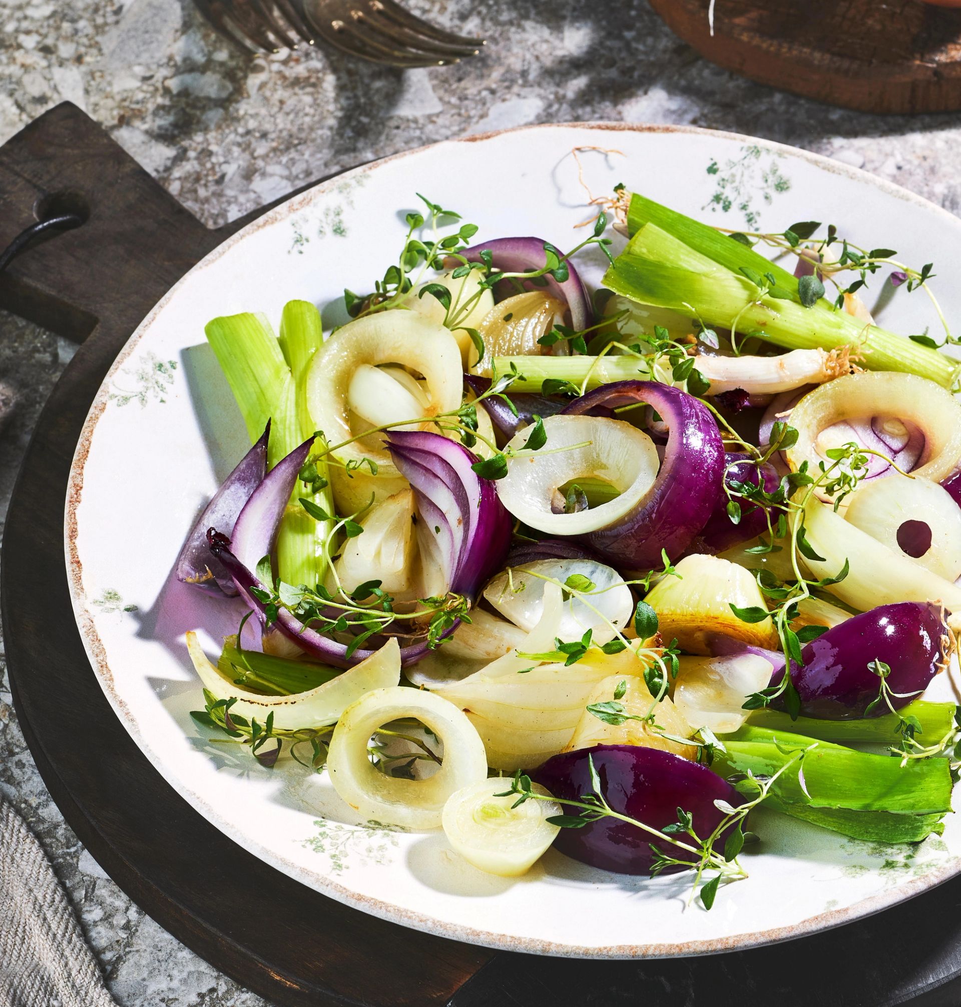 En tallerken fylt med stekte løk og vårløk i ulike farger, inkludert hvit og rød løk kuttet i ringer og båter. Retten er toppet med friske timiankvister og servert på et lyst serveringsfat.