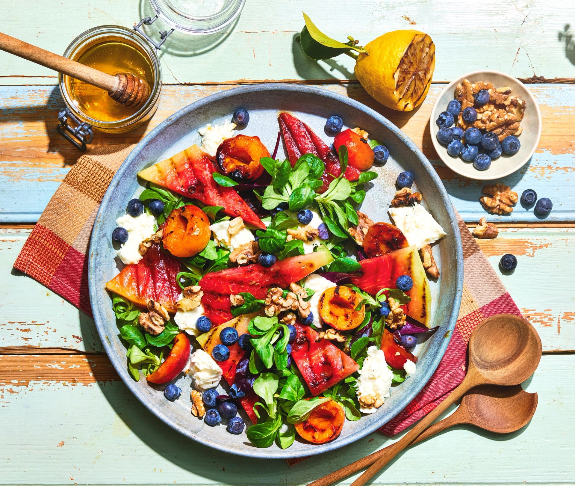 Fargerik salat i bolle sett ovenfra, med grønne blader, grillede steinfrukter, vannmelon, blåbær, valnøtter og biter av hvit ost, servert på lyst trebord med trebestikk og honning ved siden av.
