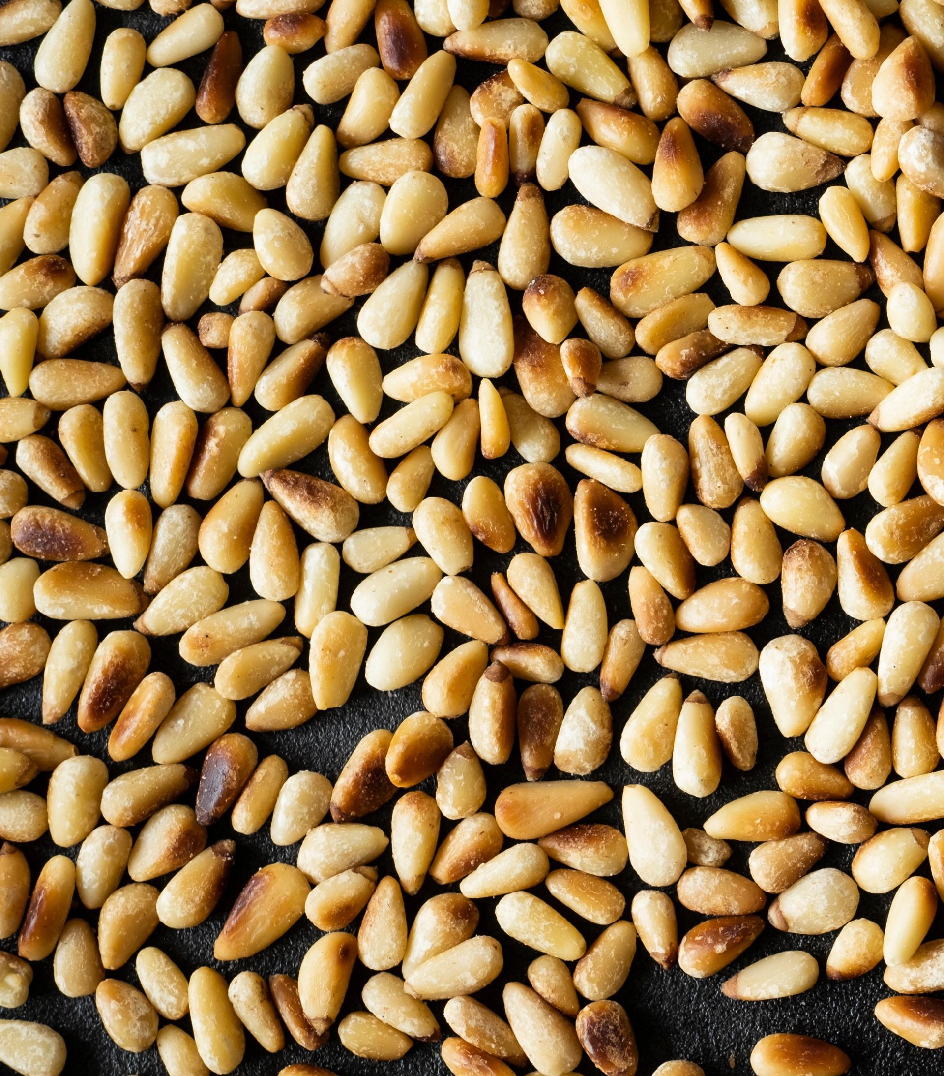 Nærbilde av mange små, avlange pinjekjerner som dekker hele bildeflaten. Kjernene varierer i farge fra lys beige til lett gyllenbrun, med enkelte litt mer ristede flekker. Overflaten virker jevn og tørr, og det er ingen andre objekter eller bakgrunnselementer synlige.