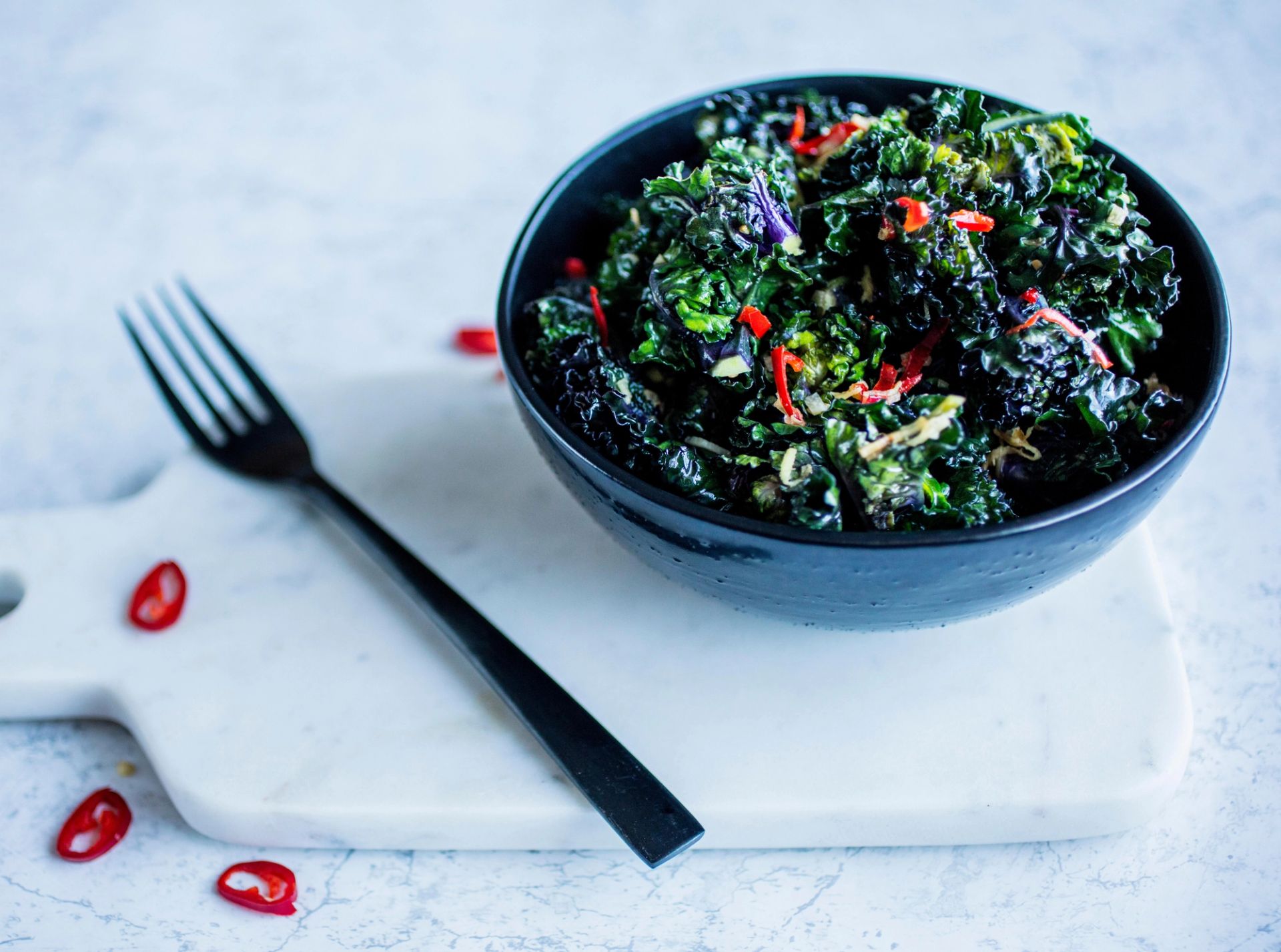En svart skål fylt med tilberedt rosettkål (flower sprouts) med røde chili‑strimler, stående på et lyst skjærebrett sammen med en gaffel og noen chili‑biter.