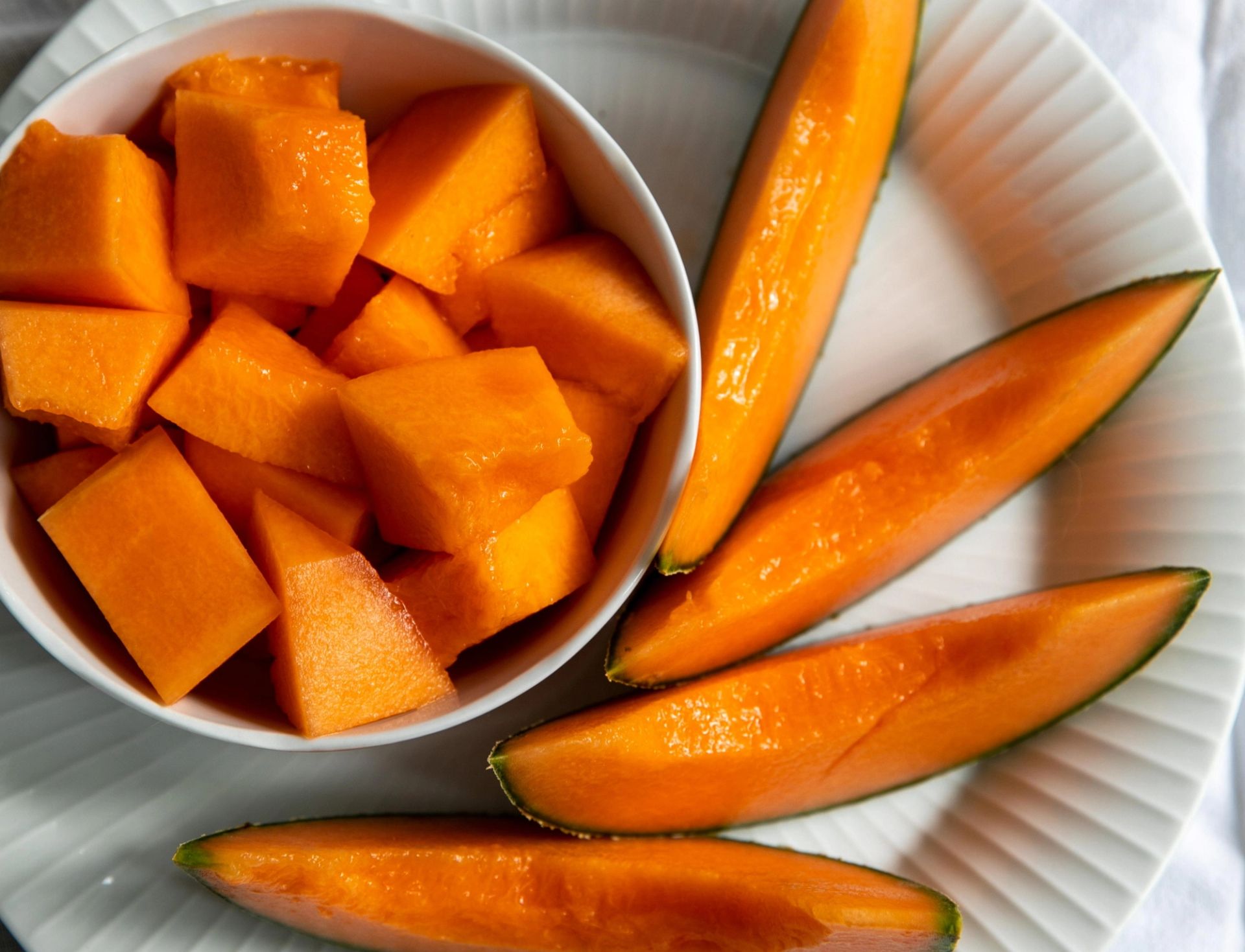 En hvit tallerken med biter av oransje melon i en skål og flere langstrakte melonskiver lagt ved siden av.