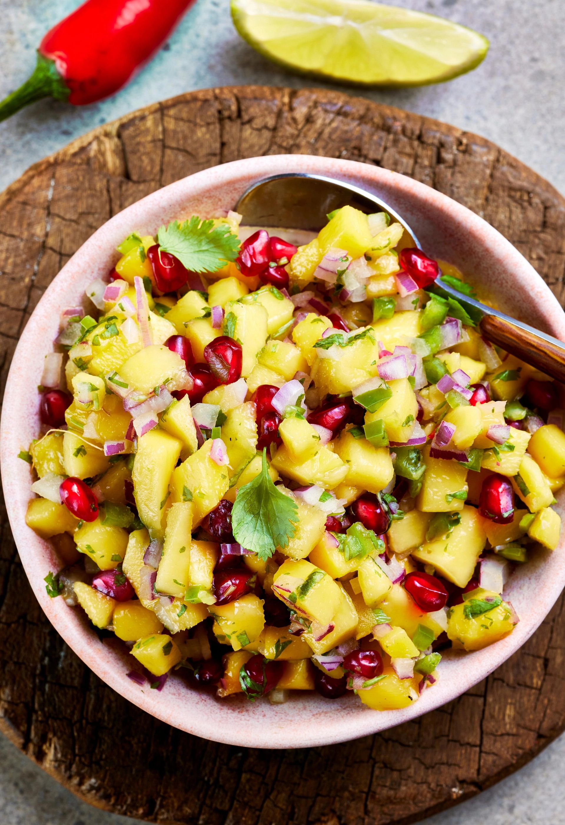 Skål med frisk salsa av mango og avokado toppet med granateplekjerner og urter, servert med skje på et rustikt trebrett, med chili og lime i bakgrunnen.