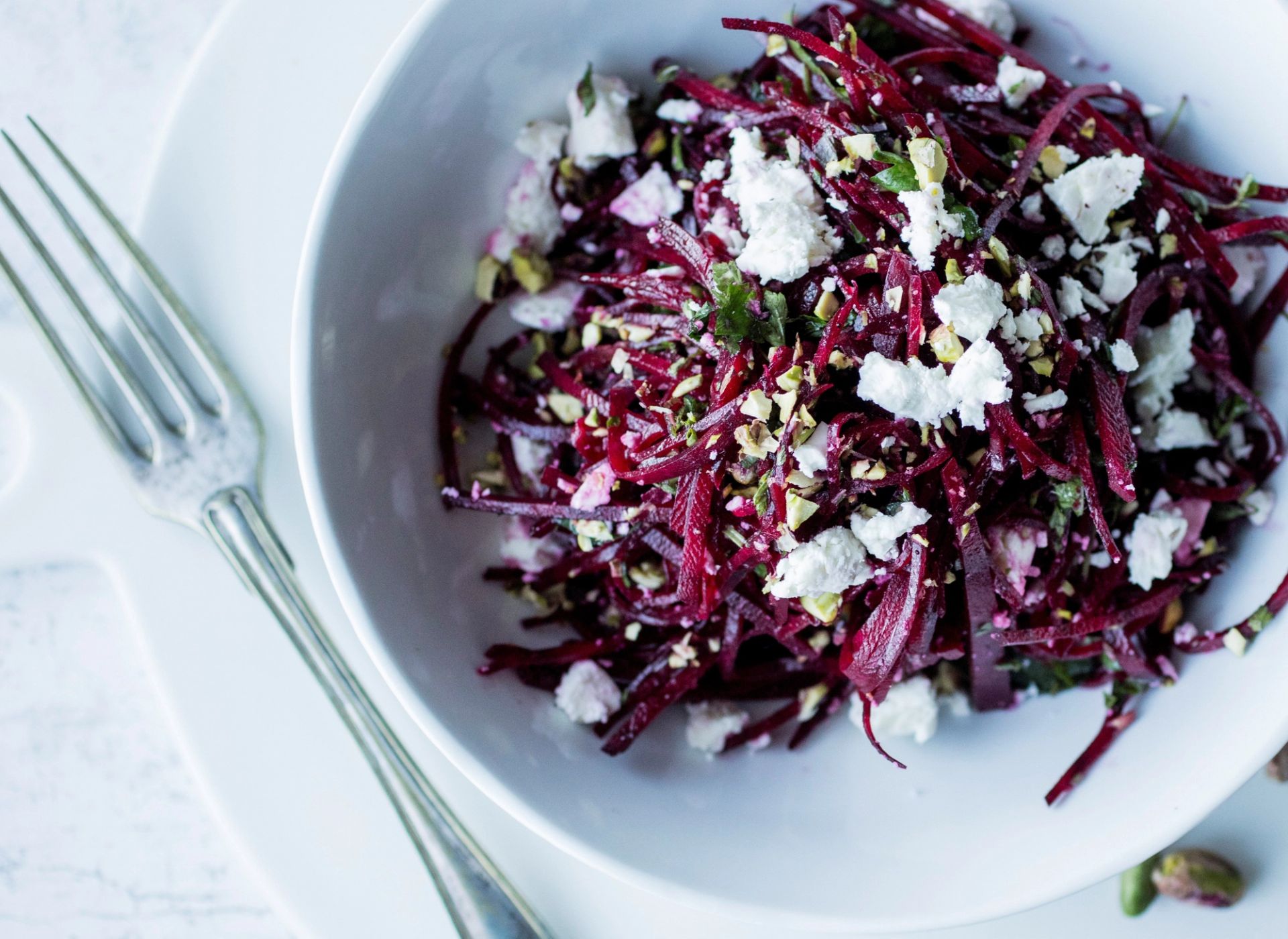 En fargerik salat av revet rødbete servert i en hvit bolle på en hvit tallerken. Salaten er toppet med smuldret fetaost og hakkede pistasjnøtter. En sølvgaffel ligger plassert ved siden av bollen.