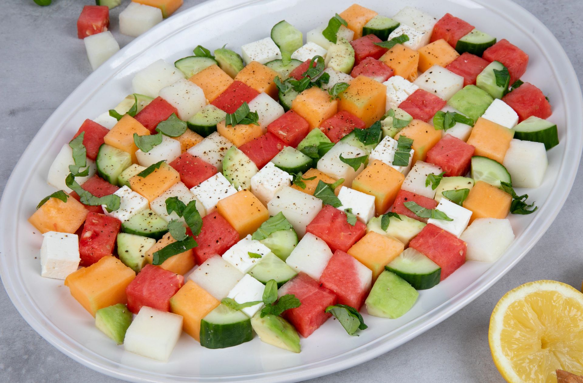 Fruktsalat med terninger av vannmelon, honningmelon, cantaloupe, agurk og fetaost, toppet med urter og pepper, servert på fat.