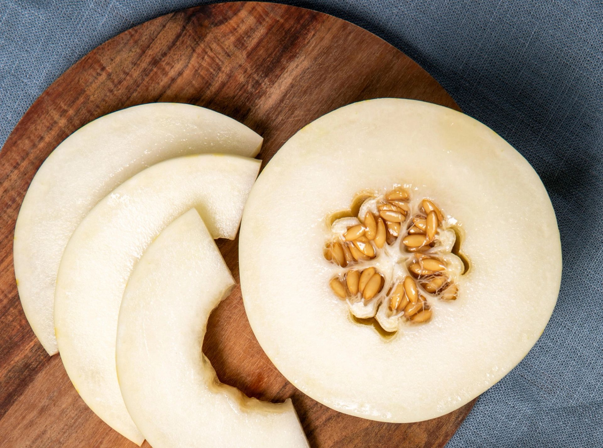 Et nærbilde av en snowball-melon på en trefjøl, der én halvdel og flere skiver ligger. Melonens lyse, nesten hvite fruktkjøtt er tydelig, og kjernene er synlige i midten av den halve melonen.