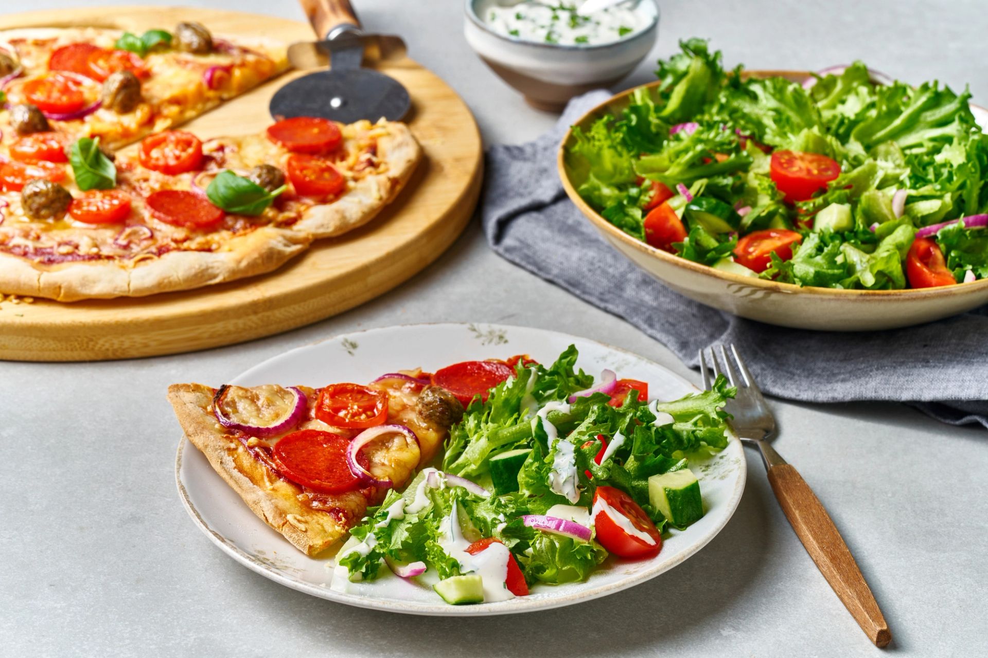 En middagstallerken med en slice hjemmelaget pizza toppet med pepperoni, tomat og rødløk, servert ved siden av en frisk grønn salat med cherrytomater, agurk og en kremet dressing.