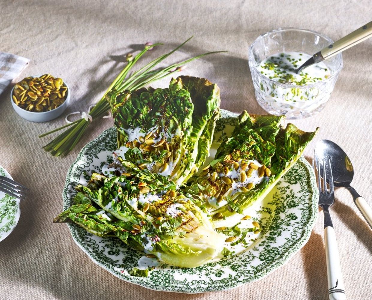 Grillet hjertesalat med synlige grillmerker, toppet med ristede gresskarkjerner, urter og en kremet dressing, servert på en grønn- og hvitmønstret tallerken. På venstre side ligger gresskarkjerner i en liten skål og en bunt gressløk. På høyre side står en glasskål med hvit dressing og en skje..