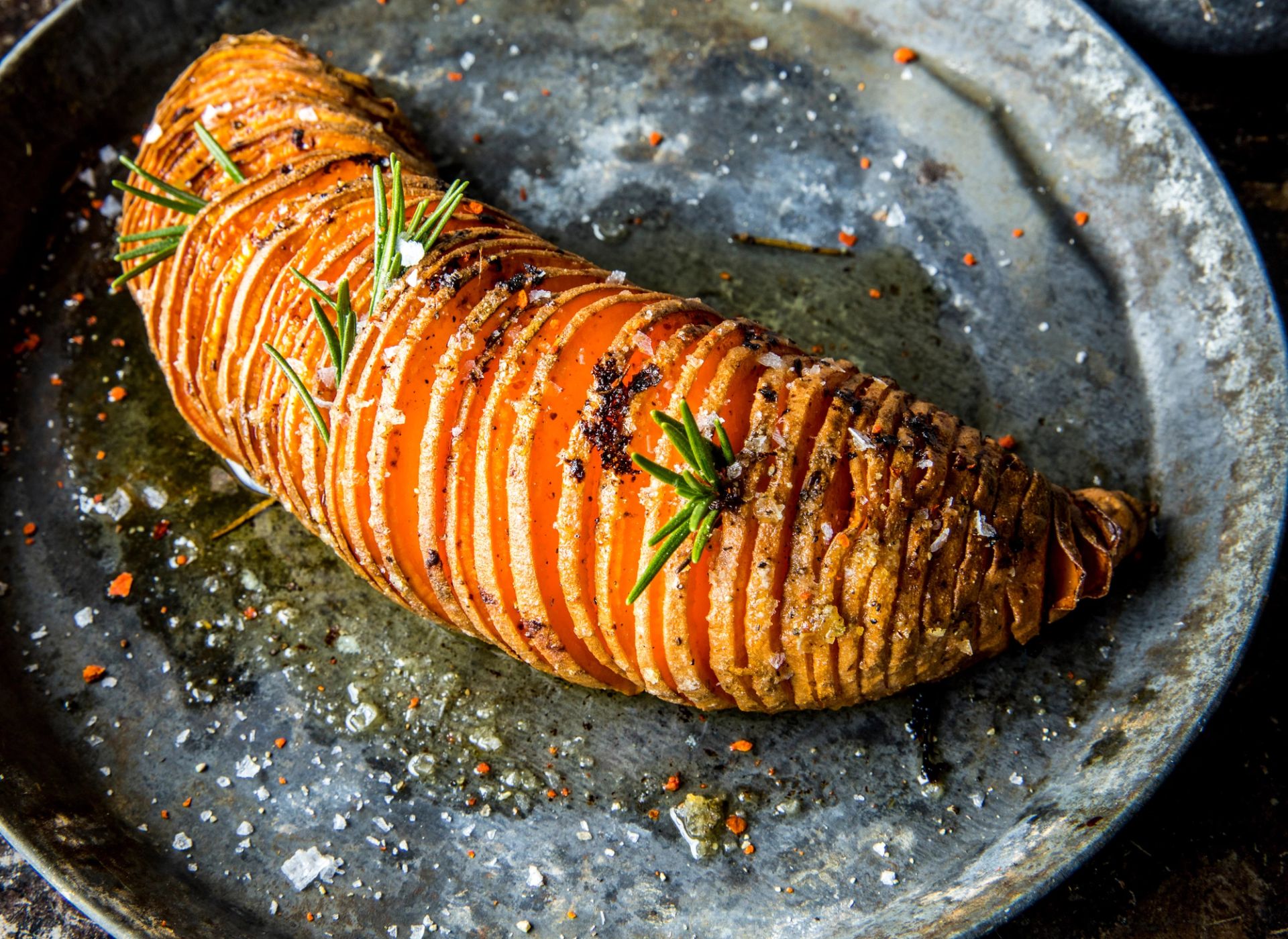 En ovnsbakt hasselback-søtpotet på et rustikt fat. Søtpoteten er skåret i tynne skiver som fortsatt henger sammen i bunnen, toppet med friske rosmarinkvaster, grovt flaksalt og kvernet pepper.