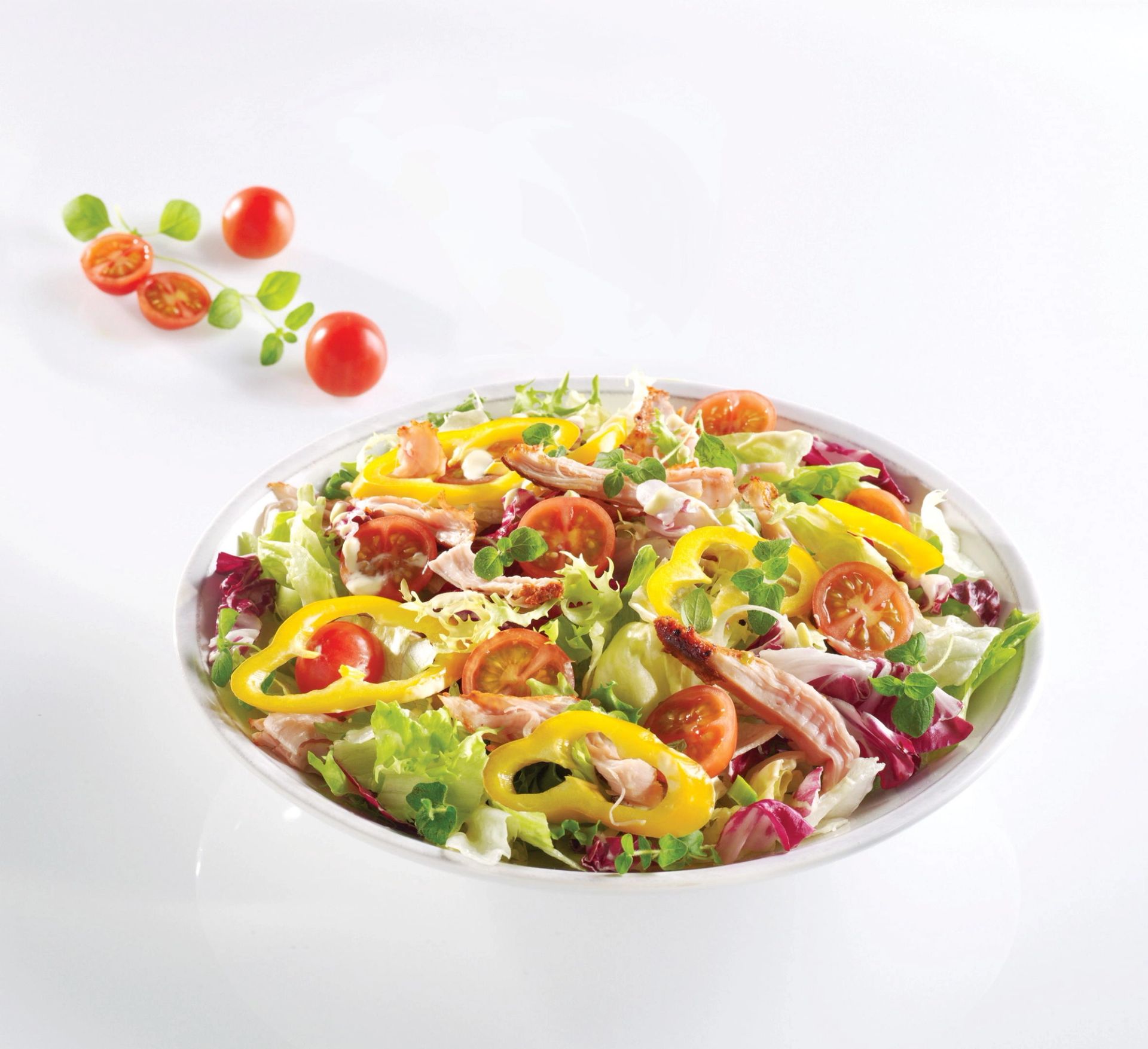 Fargerik salat med blandede salatblader, cherrytomater, gule paprikaringer og strimler av grillet kylling, servert i hvit bolle.