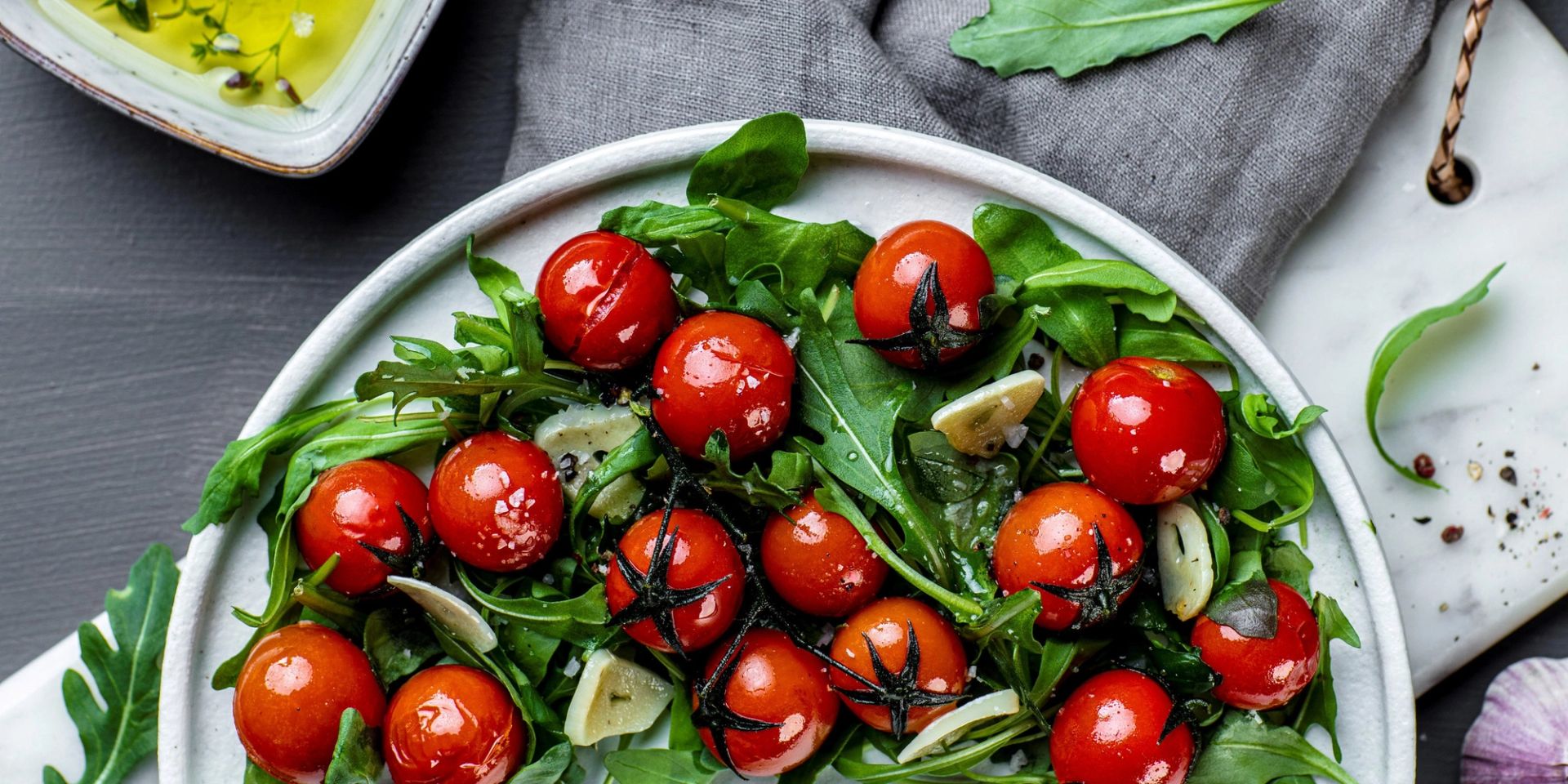 Skål med frisk salat av ruccola toppet med hele bakte cherrytomater på stilk og tynne skiver hvitløk, lett dresset med olje.