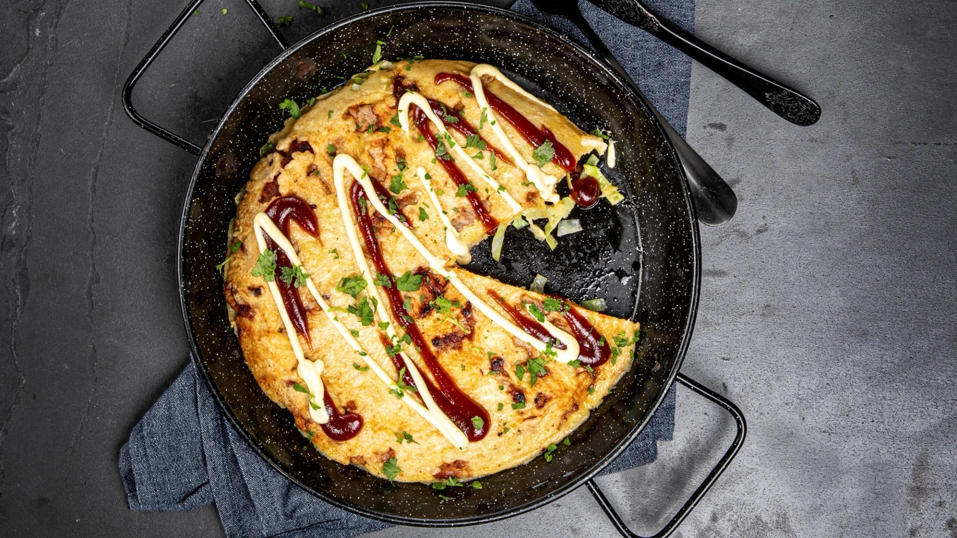 Luftig kålpannekake i stekepanne, toppet med striper av BBQ-saus og majones samt frisk persille, med et stykke skåret ut slik at fyllet inni er synlig.