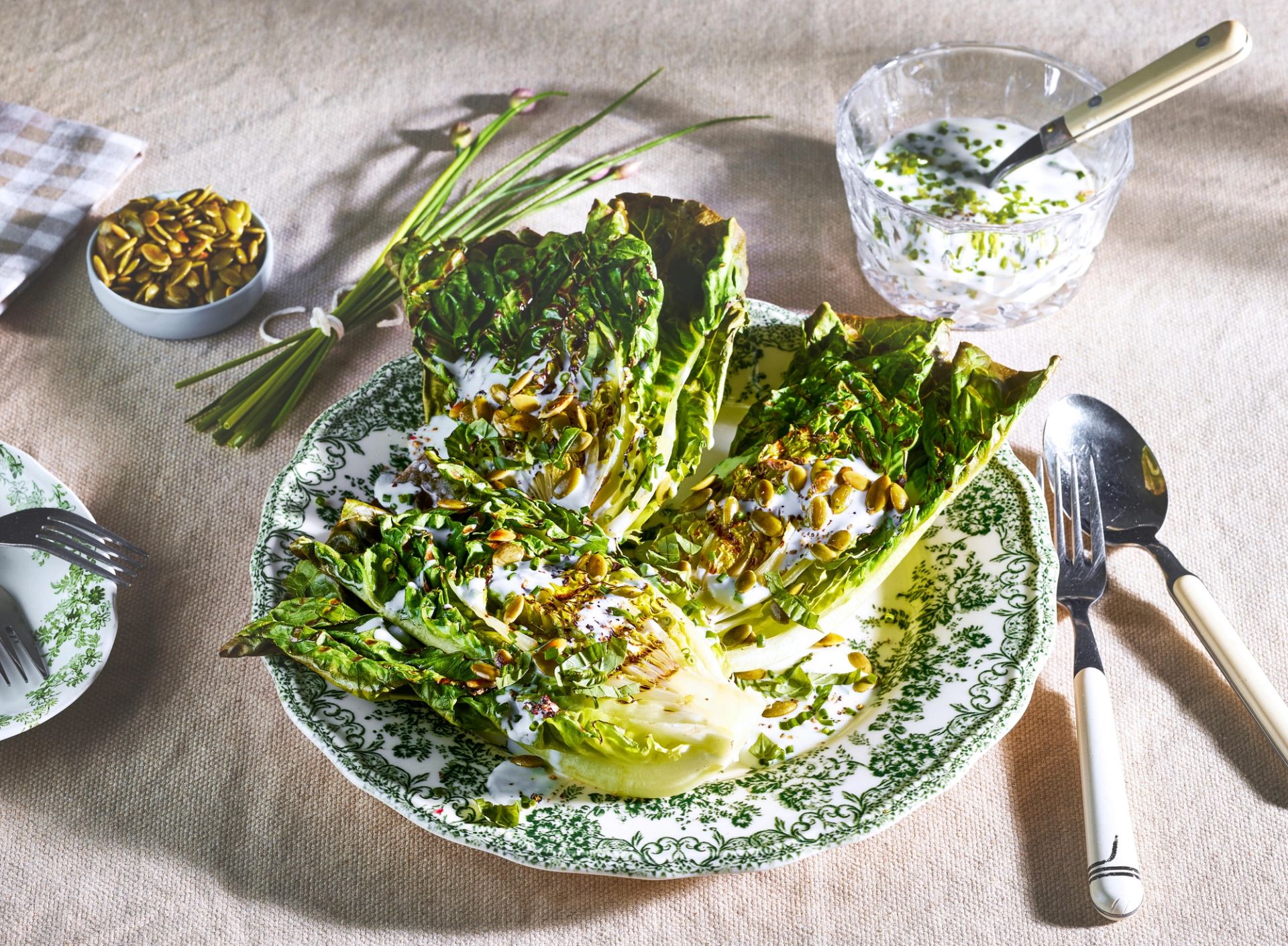 Grillet hjertesalat servert på dekorert tallerken, toppet med kremet dressing, urter og ristede frø, med bestikk og små skåler på et lyst lintøy.