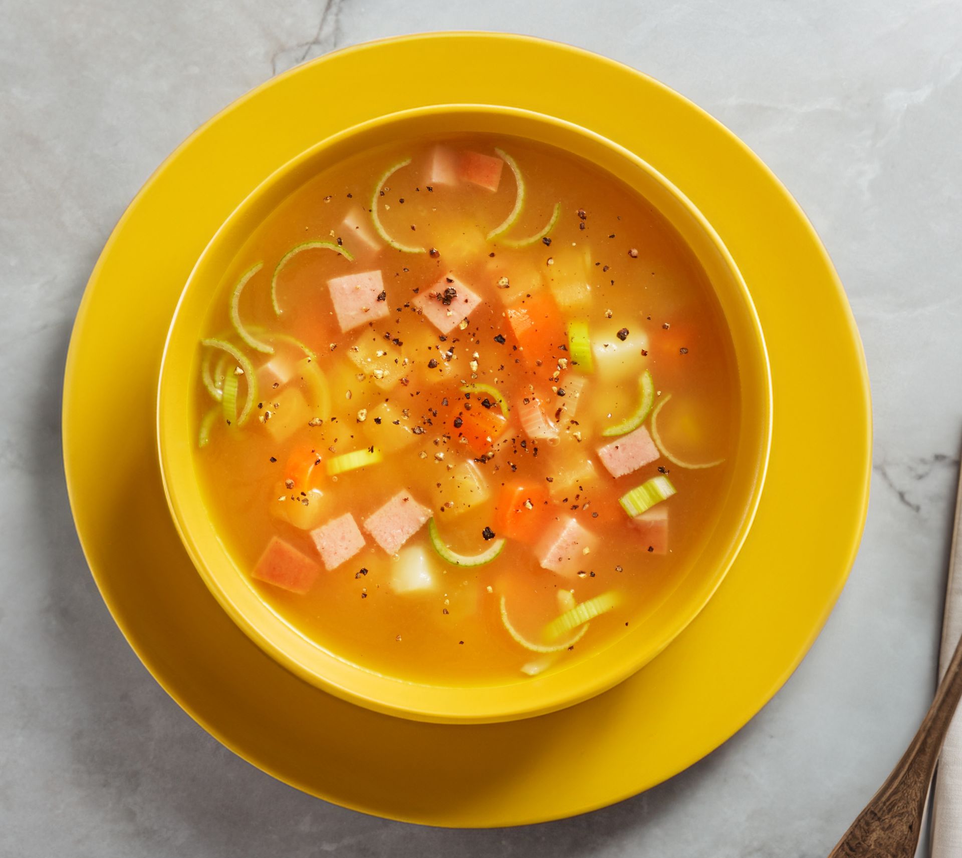 En skål med klar grønnsakssuppe på en gul tallerken, fylt med biter av gulrot, potet, purre og kjøttpølse, toppet med et dryss sort pepper, servert på et lyst underlag med en skje ved siden av.