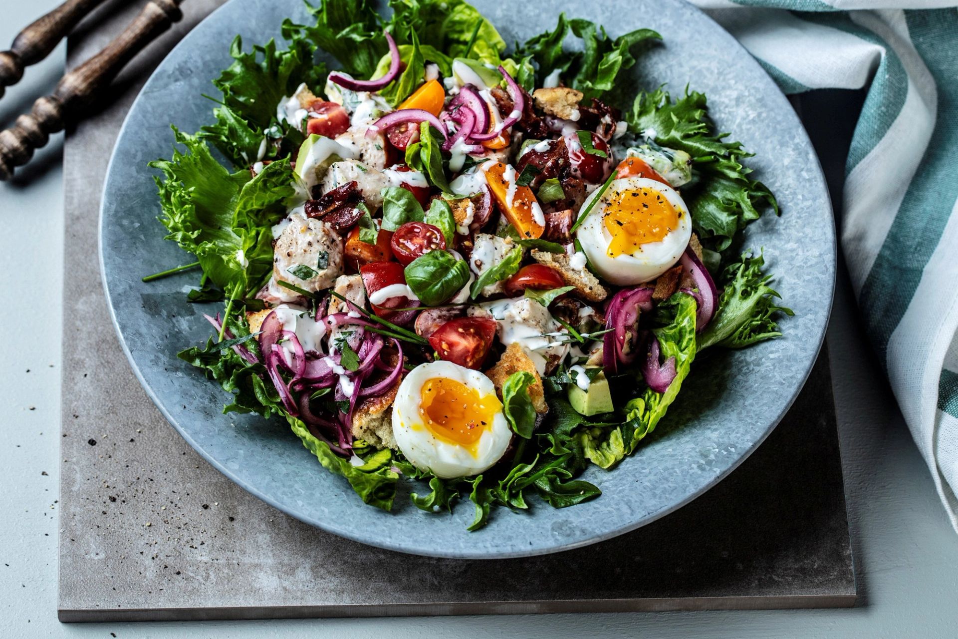 Tallerken med grønn salat sett ovenfra, toppet med delte bløtkokte egg, tomater, rødløk, sprø krutonger og biter av kylling, servert med kremet dressing på grått fat.