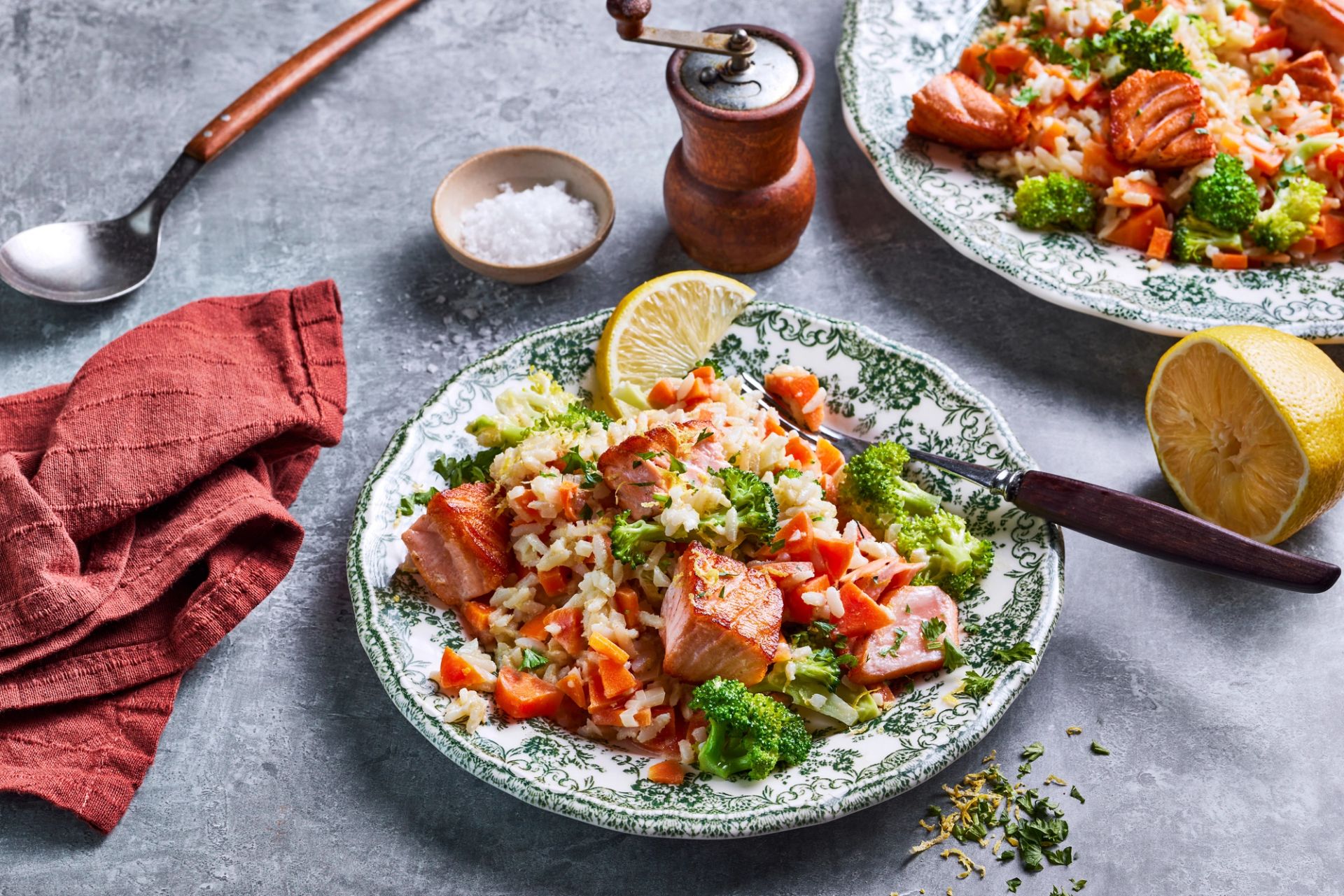 En dekorert porselenstallerken med stekt laks i biter, fullkornrisotto, brokkoli og gulrøtter. Retten er pyntet med en sitronbåt og friske urter.