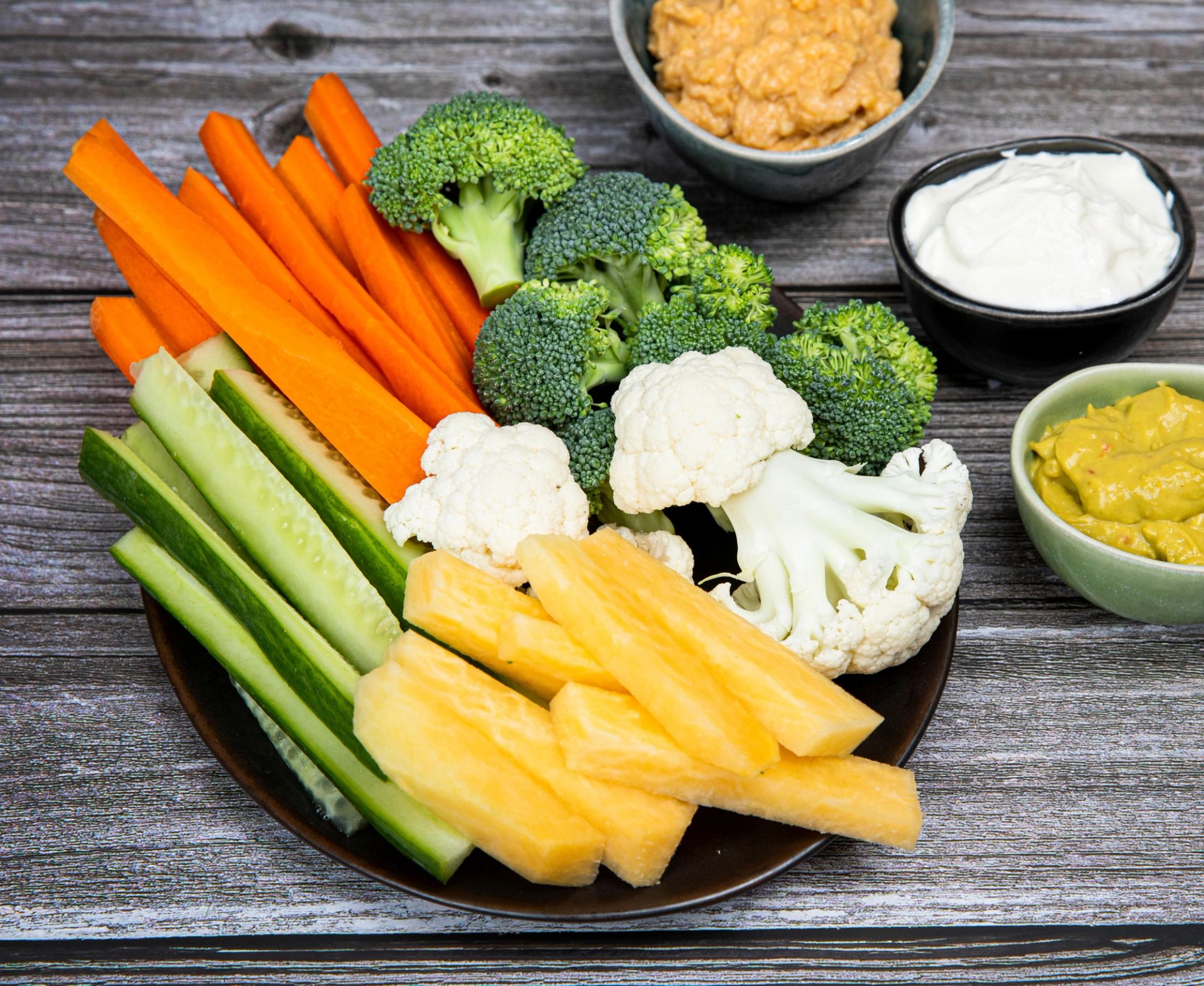 Fat med oppskårne grønnsaker og ananas, blant annet gulrotstaver, agurk, brokkoli og blomkål, servert sammen med flere skåler dip. De fargerike råvarene er pent arrangert på et mørkt fat på et rustikt trebord og fremstår som et friskt og innbydende snack- eller serveringsfat.