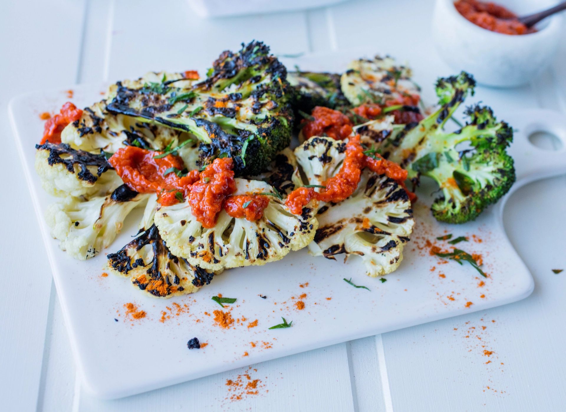 Grillet blomkål og brokkoli med sprø grillmerker, toppet med krydret harissa, friske urter og et dryss krydder – fargerik og smakfull vegetarrett servert på hvit tallerken.
