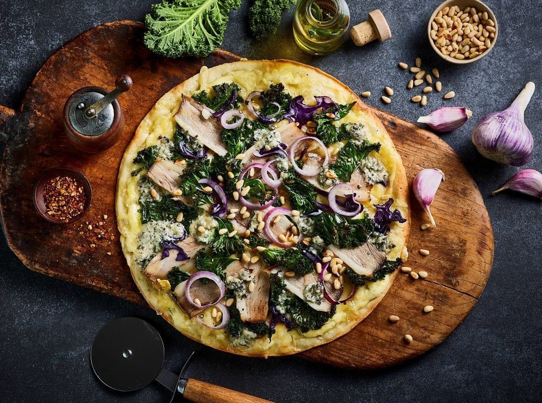 En nystekt pizza med ribbe, grønnkål, rødløk og pinjekjerner servert på en rustikk trefjøl. 
