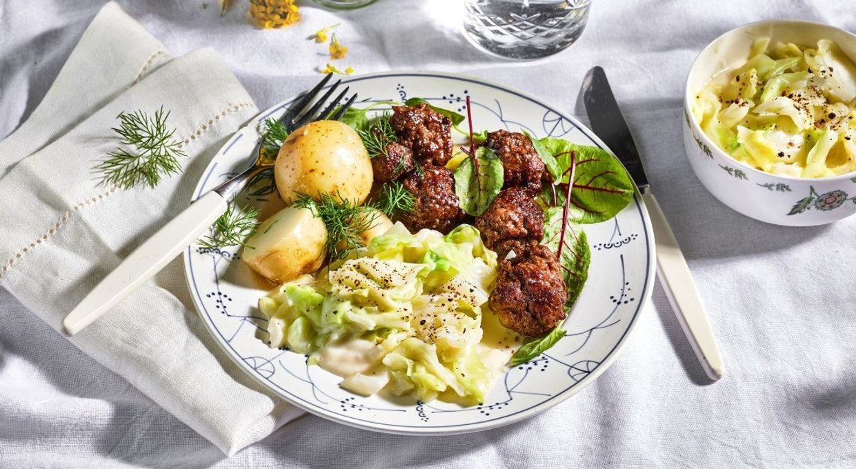 Tallerken med stuet spisskål i kremet saus servert sammen med kokte poteter og kjøttkaker, omgitt av friske urter, en bolle med mer stuet spisskål og blomster på et lyst dekket bord.