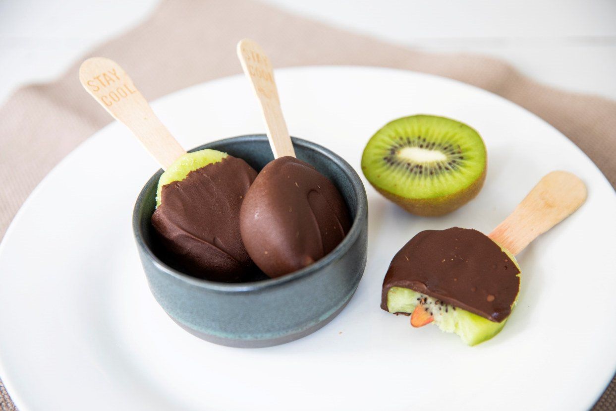 Hvit tallerken med sjokoladeovertrukne fruktis på pinne, delvis bitt av og servert i en liten skål, ved siden av en halv kiwi.