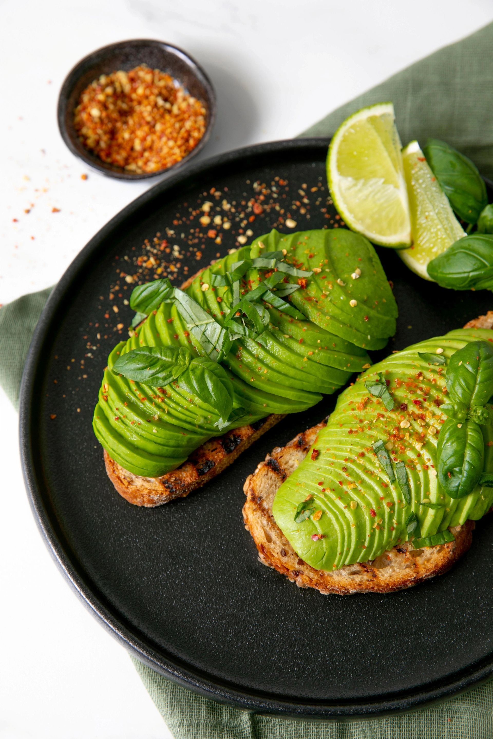 To skiver med ristet brød toppet med fine skiver av avokado, basilikum og chilikrydder. Brødskivene ligger på en mørk tallerken med noen båter lime og noen basilikumblader.
