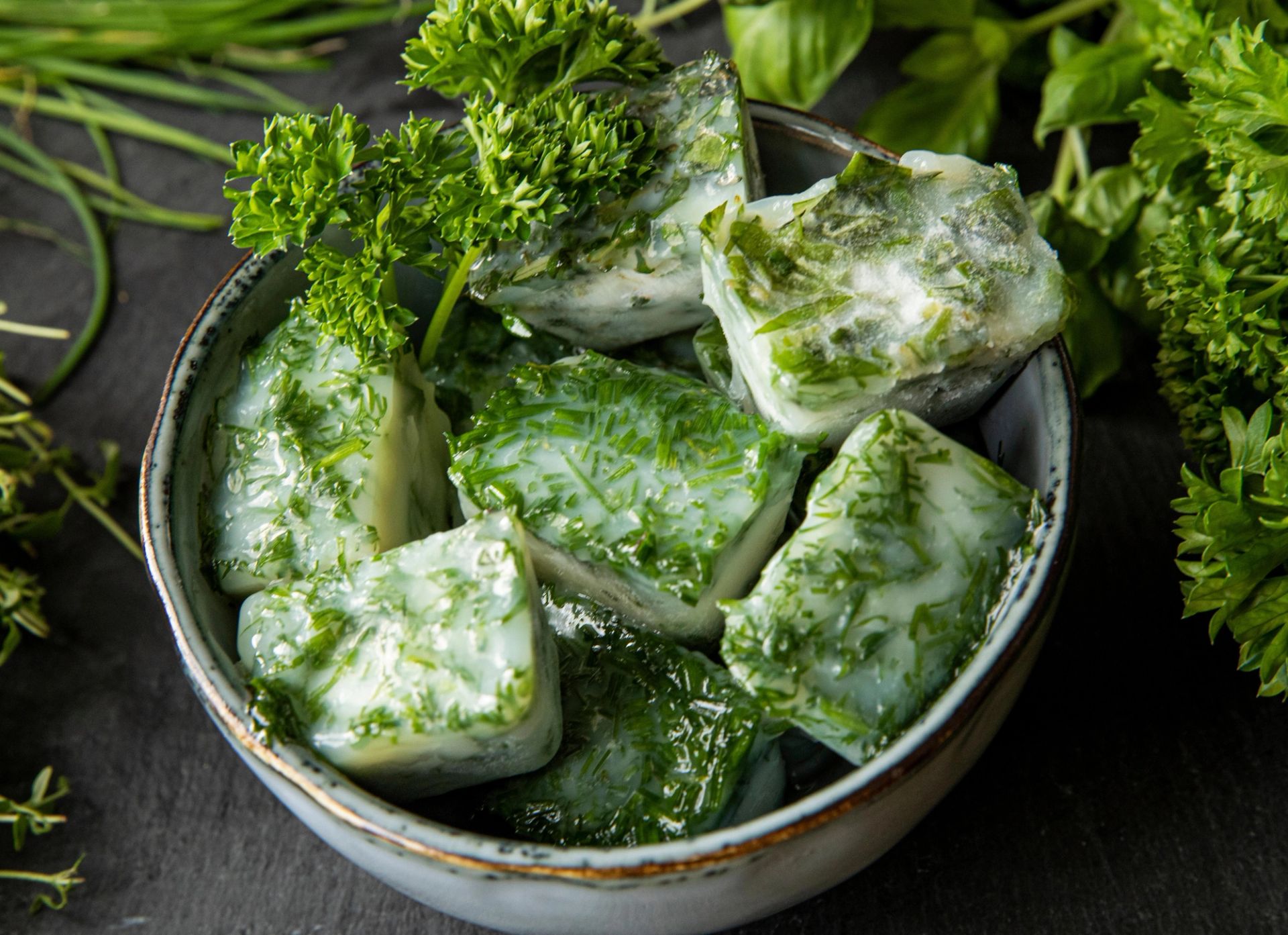 Skål med frosne smørterninger blandet med finhakket grønt krydder, som persille og urter, sett ovenfra på mørk bakgrunn med ferske urter rundt.