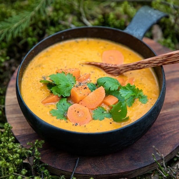 En gryte med kremet, gyllen gulrotsuppe toppet med gulrotbiter, frisk koriander og grovkvernet pepper, servert utendørs på et trebrett med tresleiv.