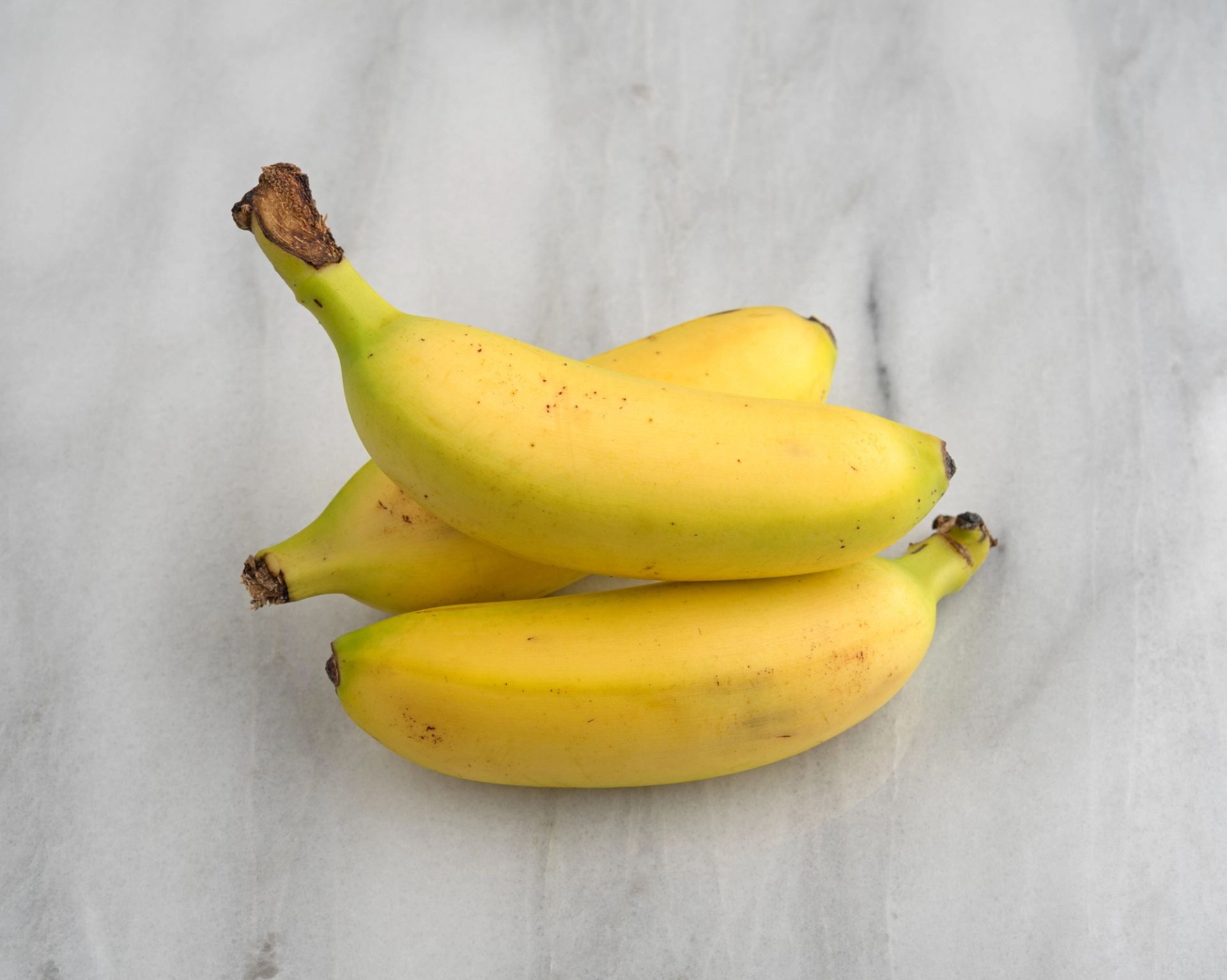 Tre modne, gule bananer som ligger stablet oppå hverandre på en lys marmorlignende overflate, sett ovenfra. Skallet har små brune prikker og stilkene er synlige.