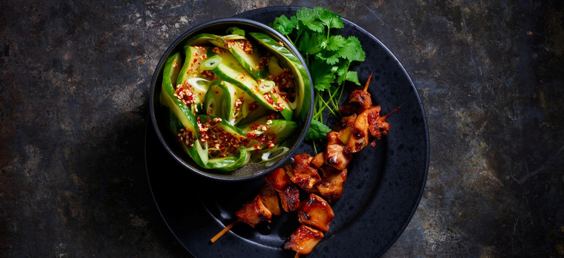 Spicy oi muchim (agurksalat) i skål med dak kkochi (kyllingspyd) servert på en mørk tallerken