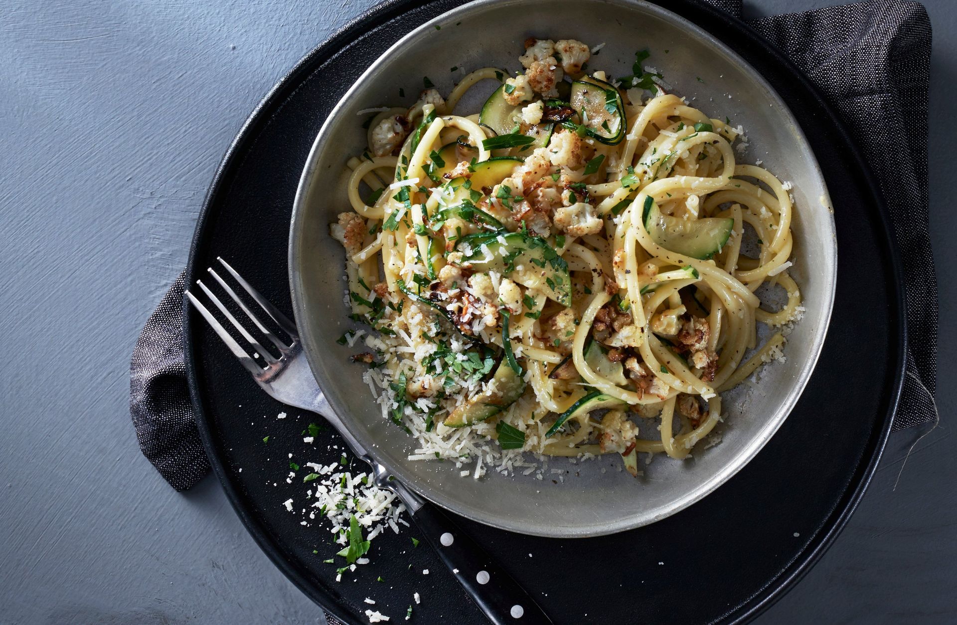 Tallerken med linguine-pasta vendt i kremet carbonarasaus, toppet med sprøstekt blomkål, urter og revet parmesan. Gaffel ligger klar.