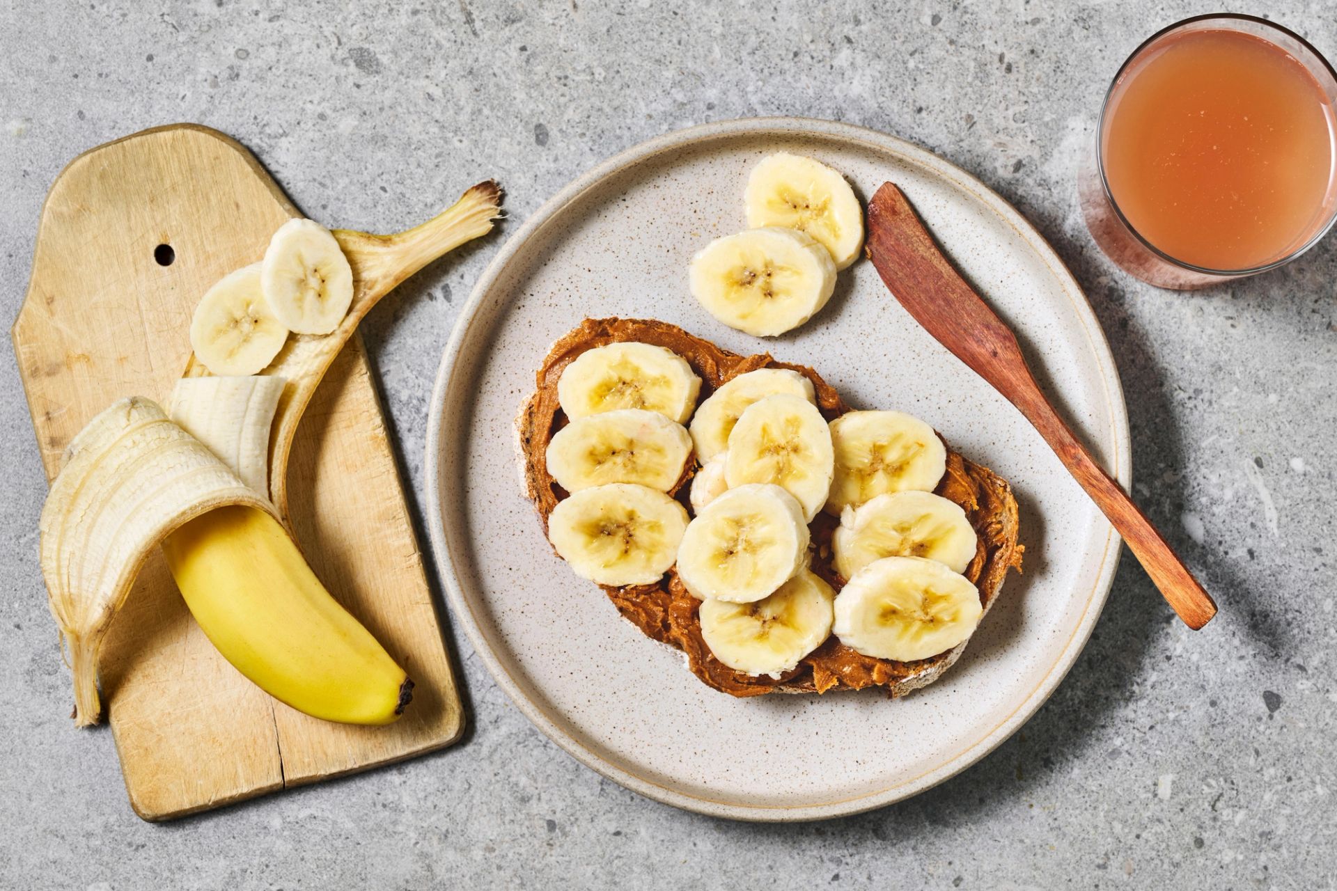 Brødskive med skivet banan og peanøtter toppet med honning, servert med hele bananer på lys bakgrunn