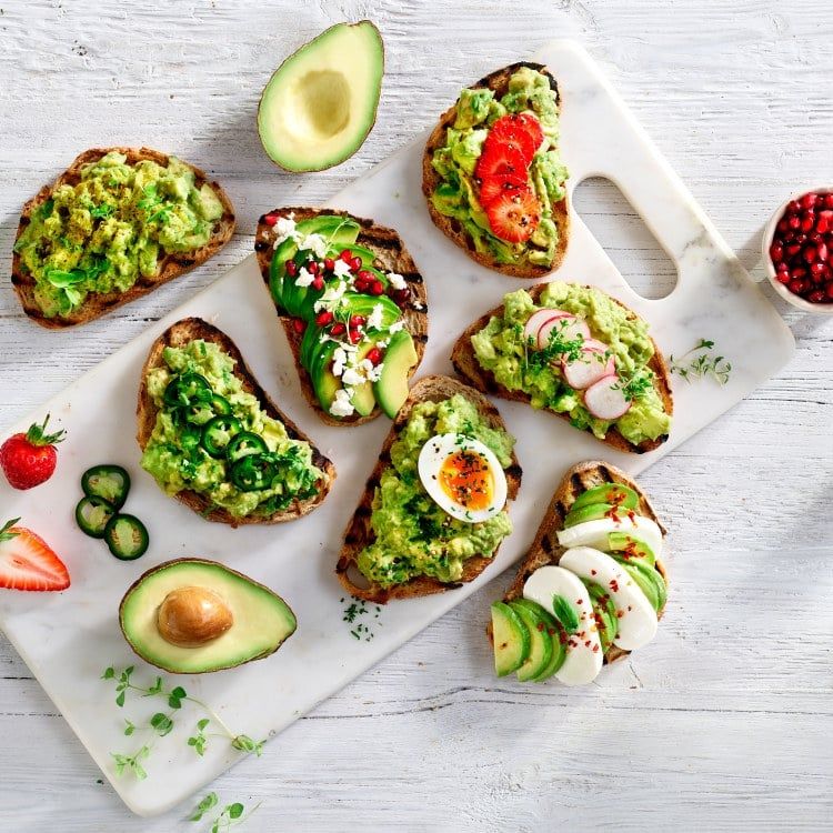 Flere skiver ristet brød toppet med skivet og most, med ulike fargerike ingredienser, inkludert jordbær, jalapeno, kokt egg, mozzarella, reddik, fetaost og granateple, arrangert på et hvitt skjærebrett med halv avokado og bær rundt.