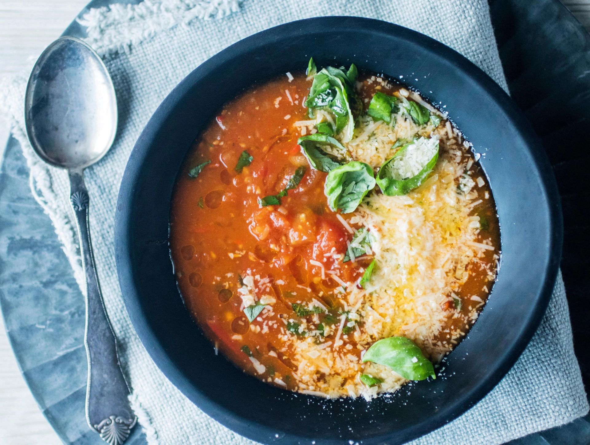 Tomatsuppe servert i en mørk skål, toppet med revet parmesan og rosenkål, med en skje ved siden av på et lyst tekstilunderlag.
