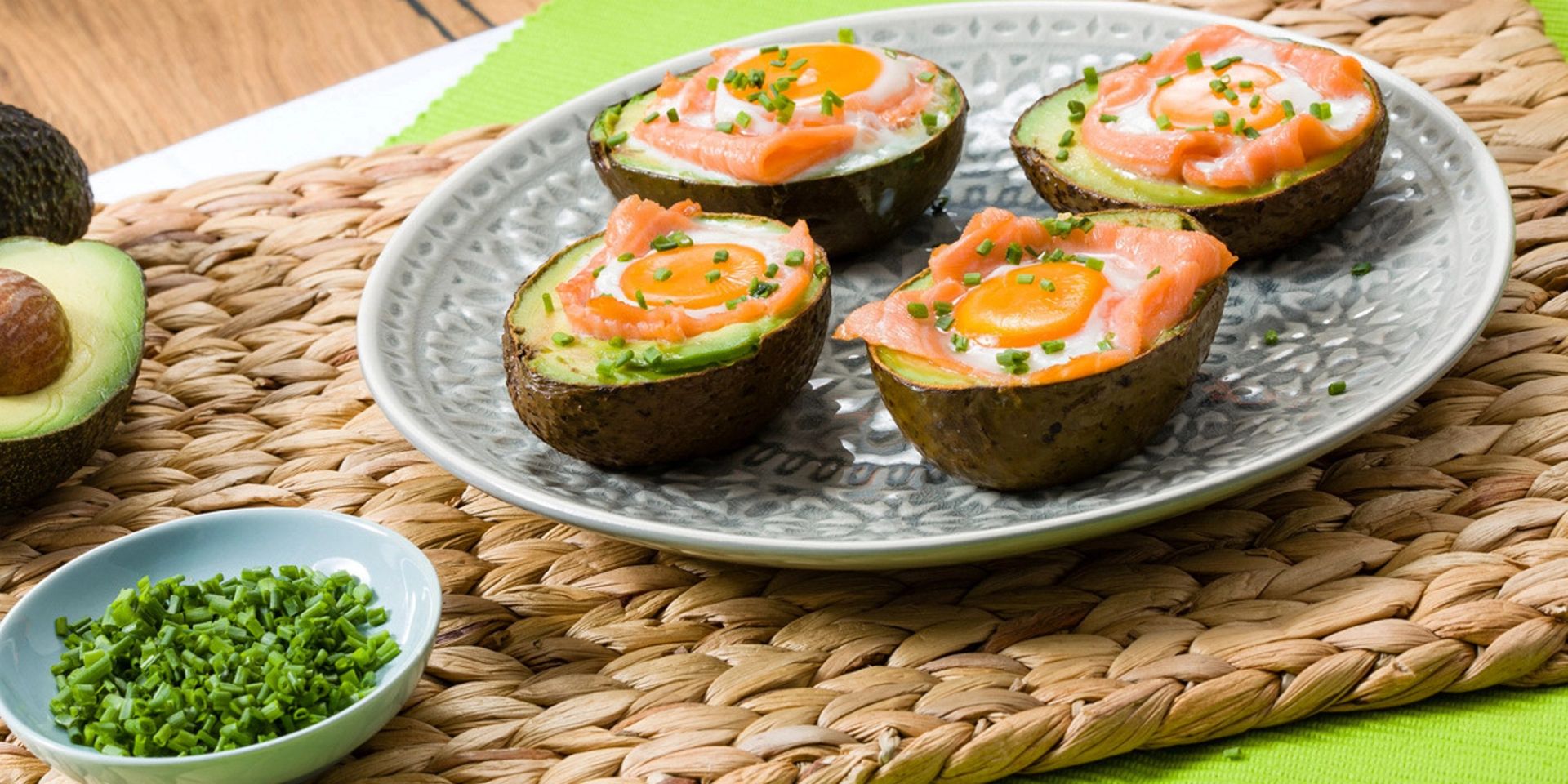 Halve avokadoer fylt med egg og skiver av røkt laks, toppet med finhakket gressløk. Servert på et mønstret fat med ekstra gressløk i en liten skål og ferske avokadoer i bakgrunnen.
