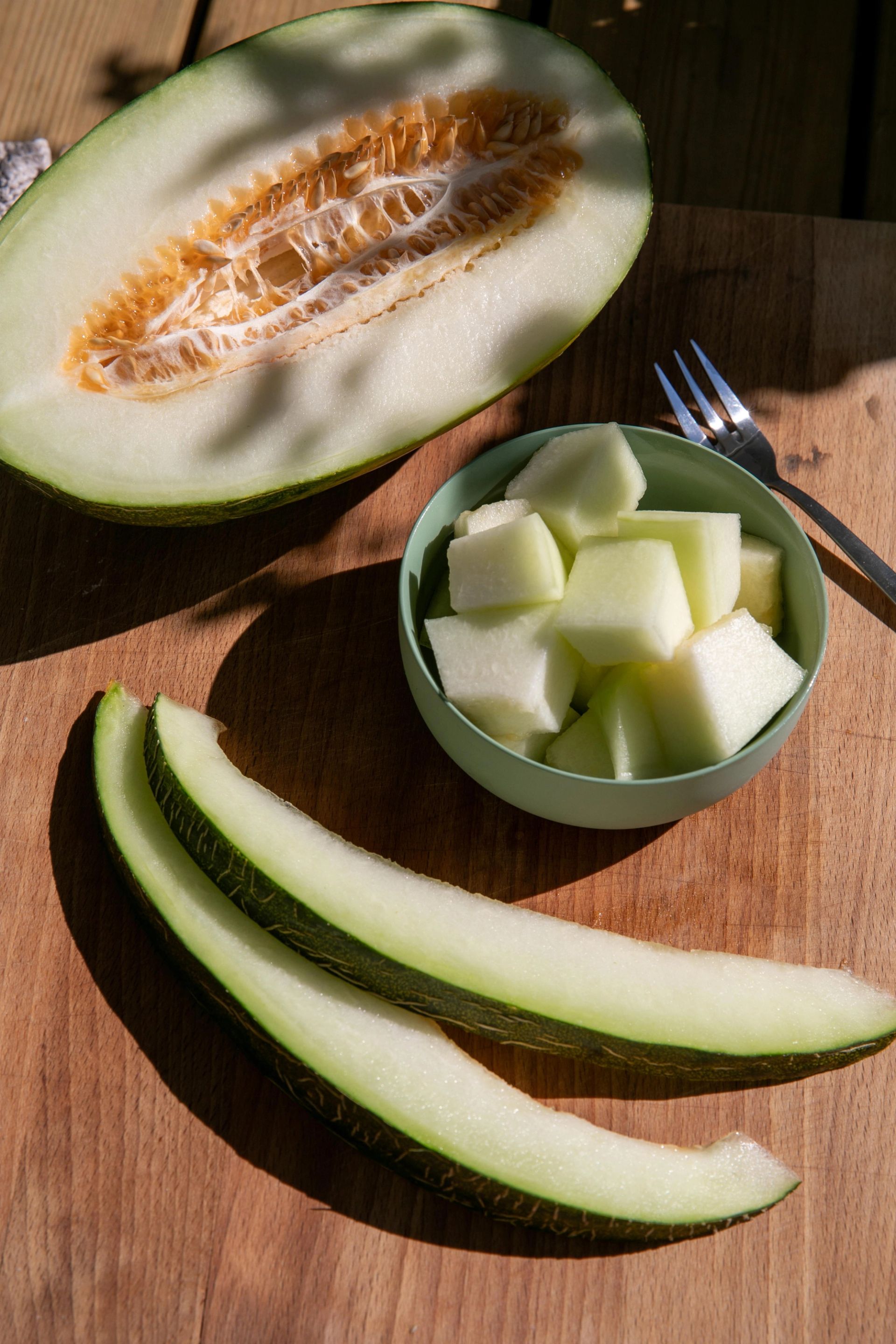 En halv lysgrønn Piel de Sapo melon med synlige frø ligger bak en liten skål fylt med terninger av melon, mens tre lange melonskiver ligger på et skjærebrett foran