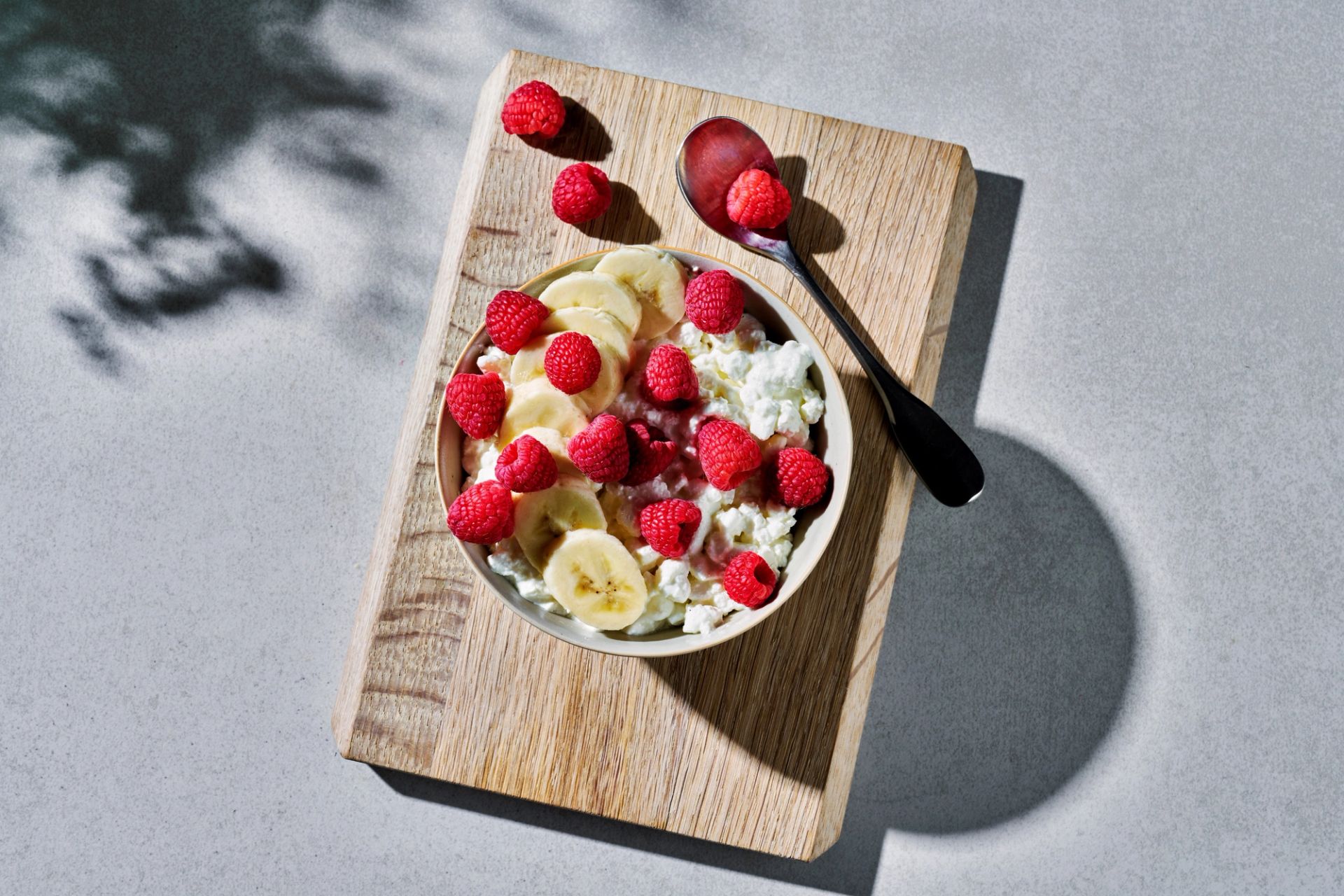 Cottage cheese i en skål toppet med skiver av banan og friske bringebær, servert på et lite trebrett med en skje ved siden av.