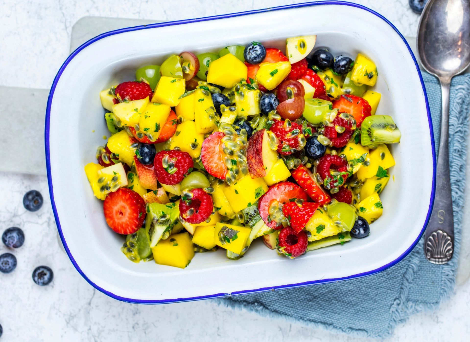 En rektangulær, hvit serveringsform fylt med frisk fruktsalat. Salaten består av terninger av gul mango, halve jordbær, blåbær, grønne druer, røde druer og små biter av en grønn frukt med svarte kjerner, alt blandet med en hakket, grønn urteblanding. Formen står på et lyst underlag med et sammenfoldet blått kjøkkenhåndkle og en metallskje ved siden av. Rundt formen ligger noen løse blåbær.