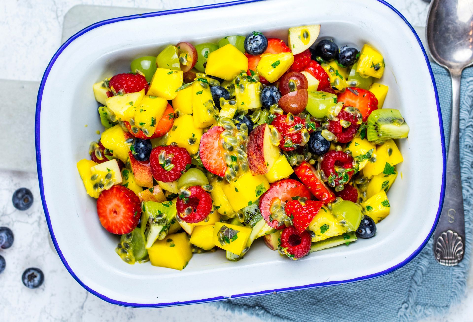 En rektangulær, hvit serveringsform fylt med frisk fruktsalat. Salaten består av terninger av gul mango, halve jordbær, blåbær, grønne druer, røde druer og små biter av en grønn frukt med svarte kjerner, alt blandet med en hakket, grønn urteblanding. Formen står på et lyst underlag med et sammenfoldet blått kjøkkenhåndkle og en metallskje ved siden av. Rundt formen ligger noen løse blåbær.
