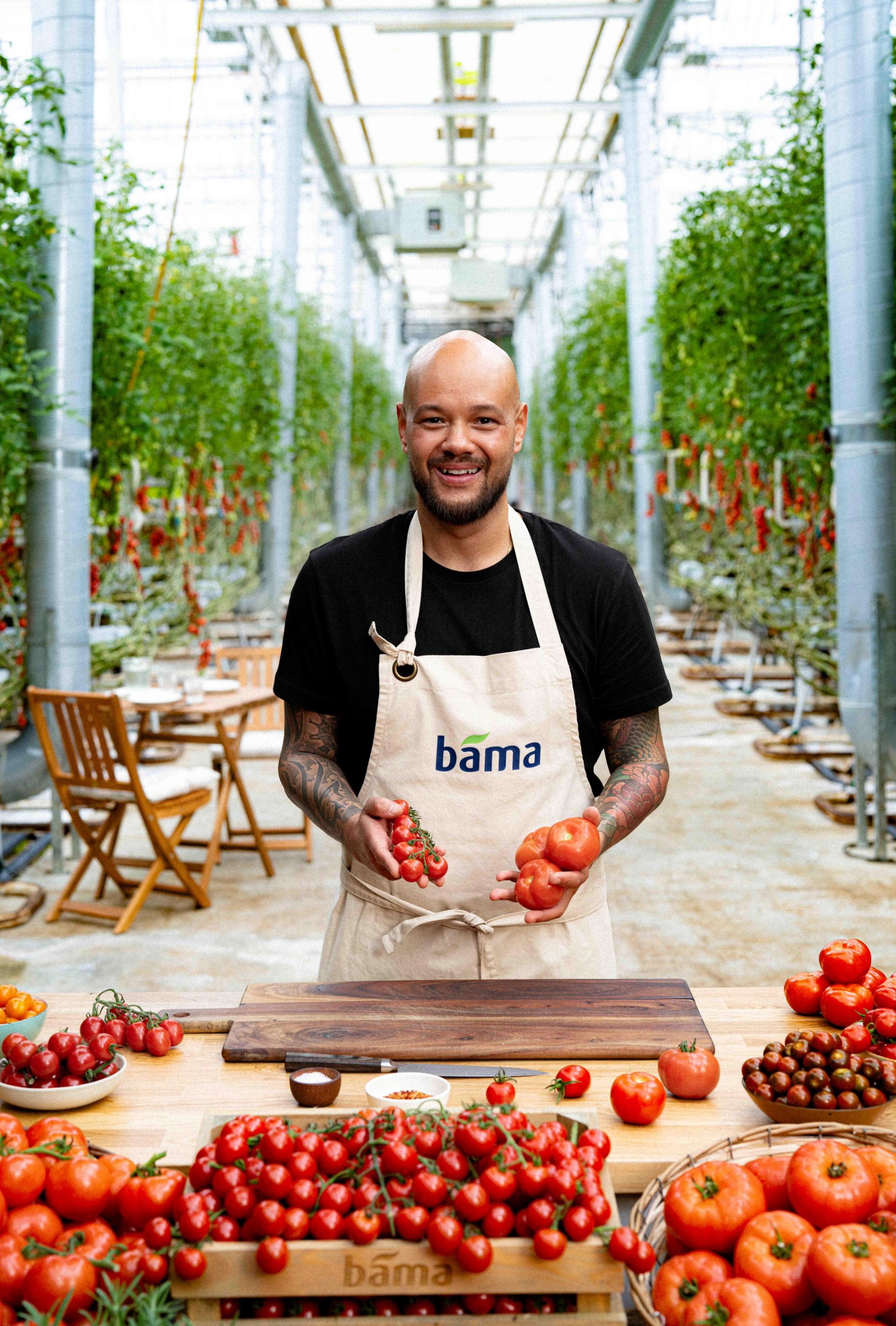 Mann i drivhus holder tomater i hendene, med BAMA-forkle og bord med ulike tomatsorter foran seg.