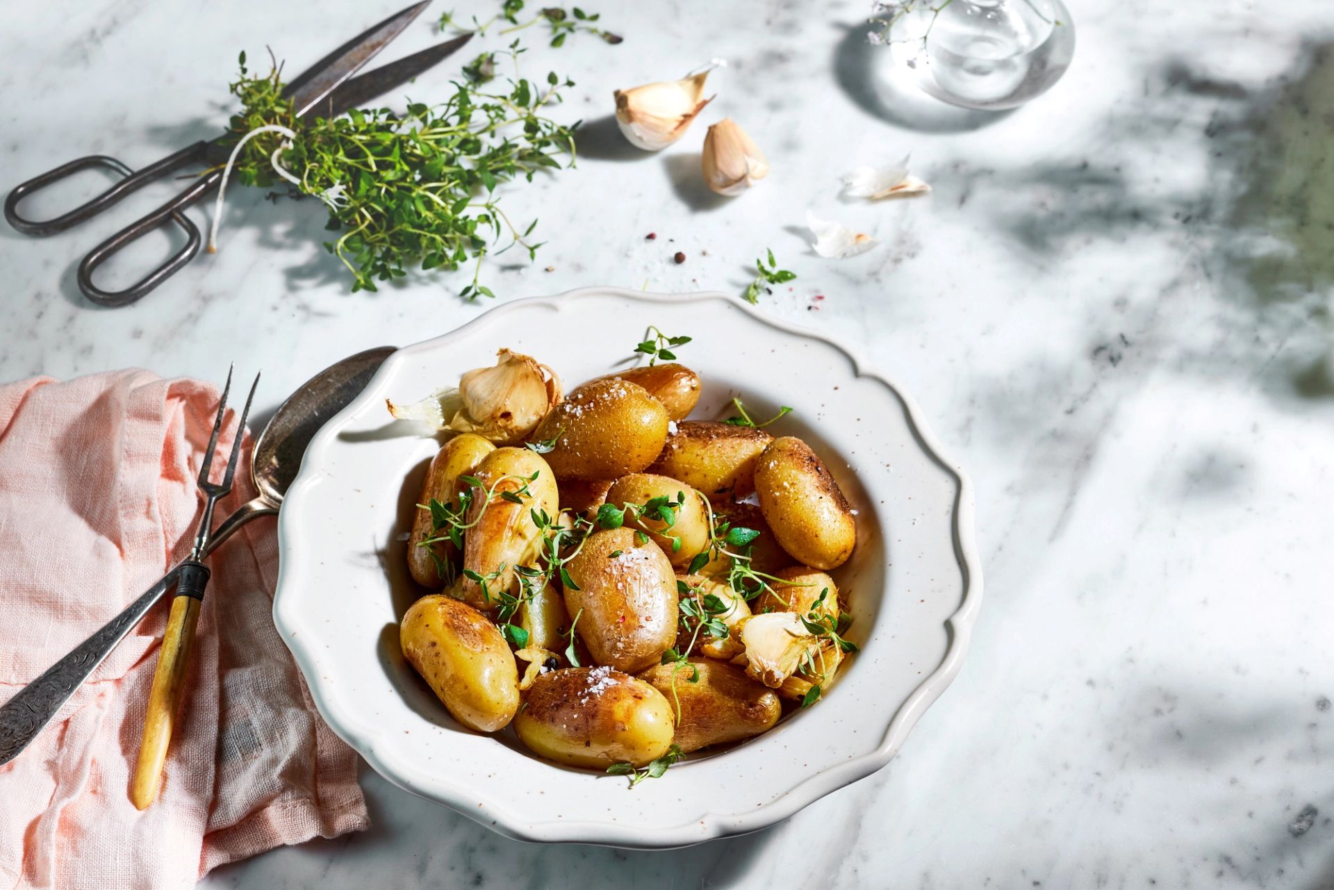 Ovnsbakte småpoteter med hele hvitløksfedd og frisk timian, servert i en hvit bolle på marmorbord.